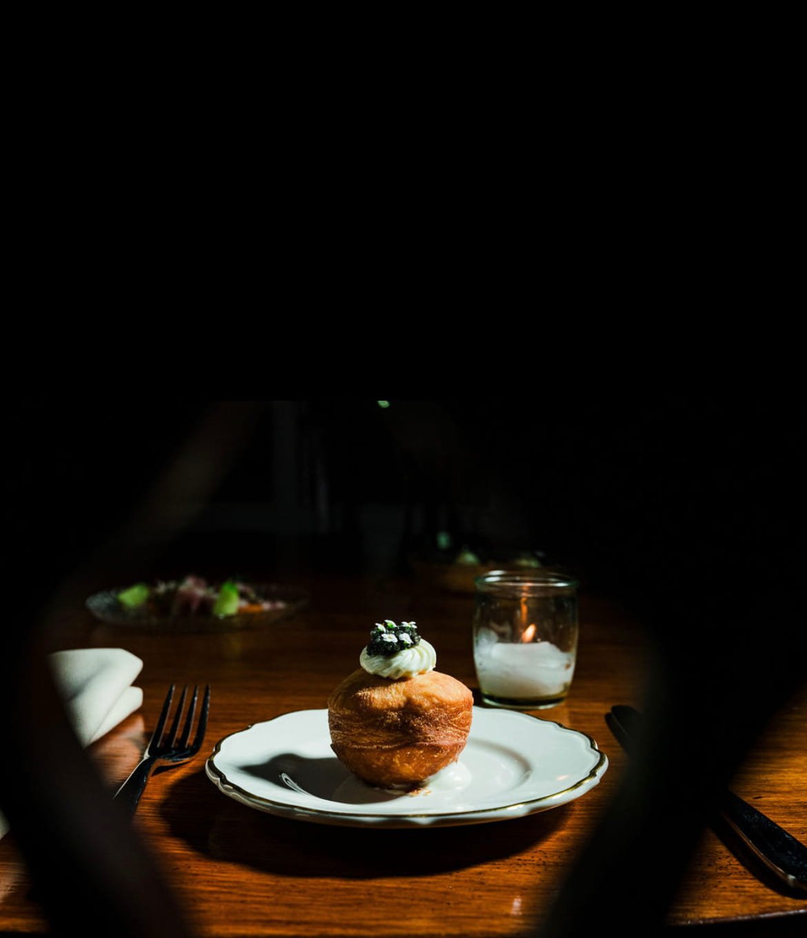 A dessert resembling a bread bowl with a dollop of cream or frosting topped with berries on a white plate, on a wooden table with a fork and knife nearby, in a dimly lit setting