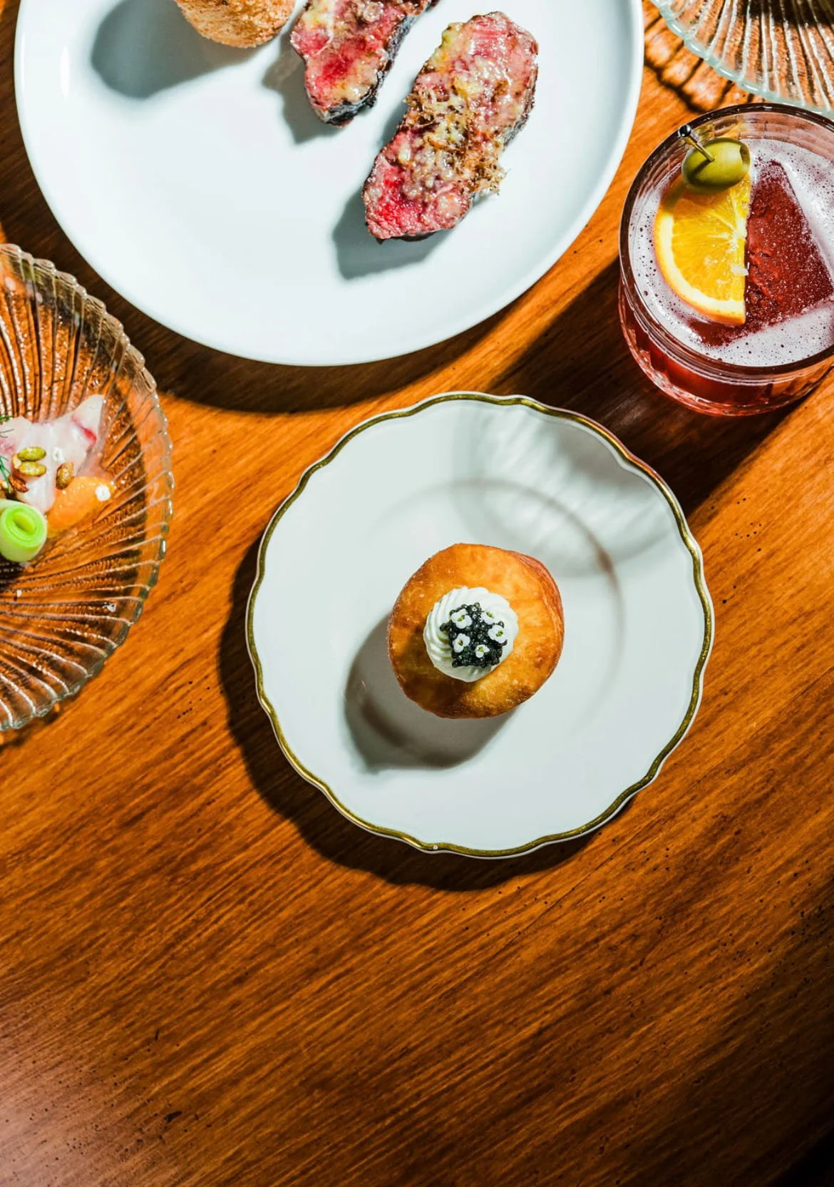 A wooden table with various fancy appetizers and a drink. There are two pieces of uncooked steak on a white plate, a cupcake with white frosting and black caviar on a decorative white plate, a glass with a red cocktail garnished with an orange slice and a green olive, and other small dishes.