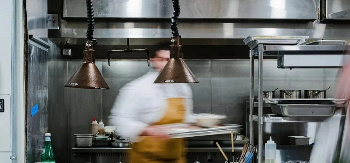 Blurred image of a chef in a professional kitchen, wearing a white coat and a yellow apron, holding a tray, with metallic kitchen equipment and utensils visible.