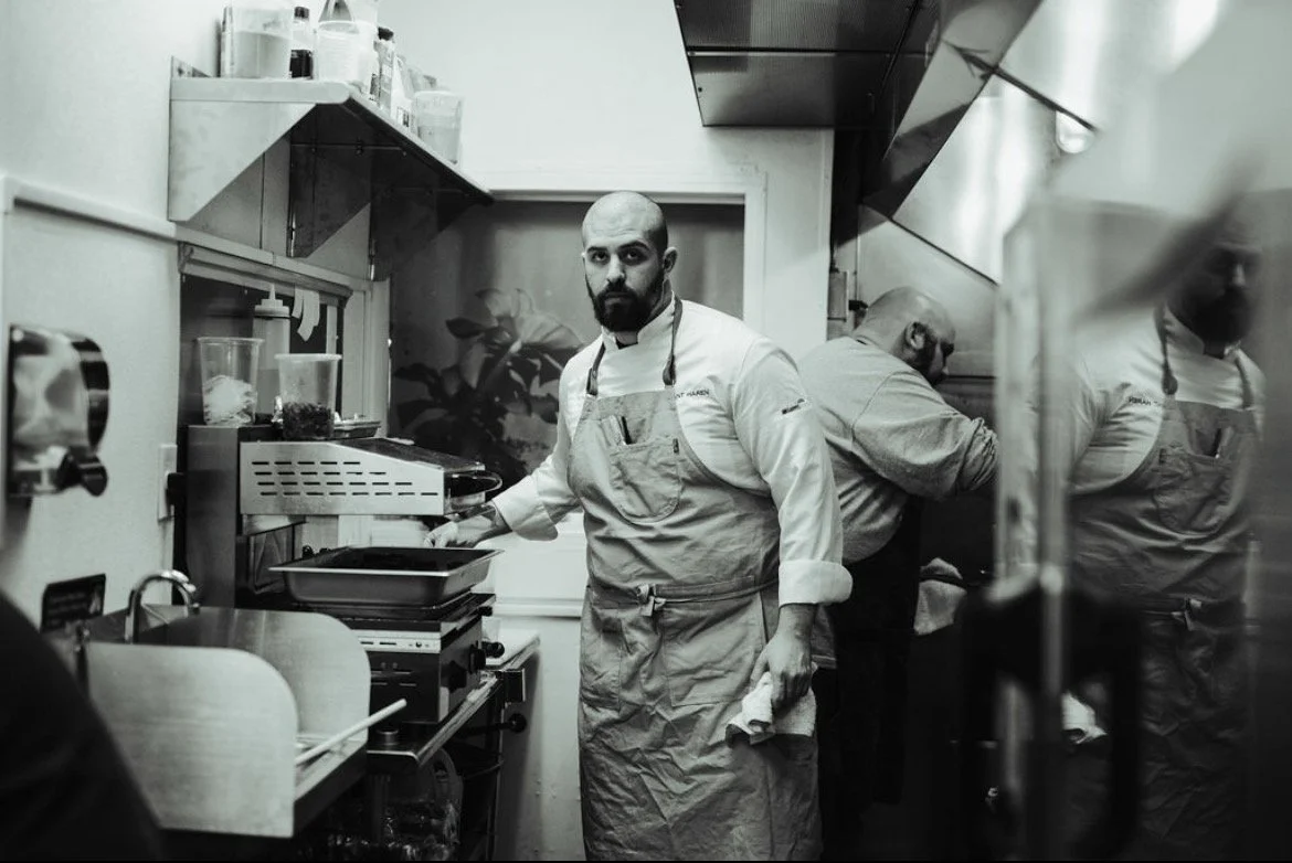 A chef in a restaurant kitchen looking at the camera, wearing a white chef's coat and apron, standing near cooking appliances with two other chefs working behind him.