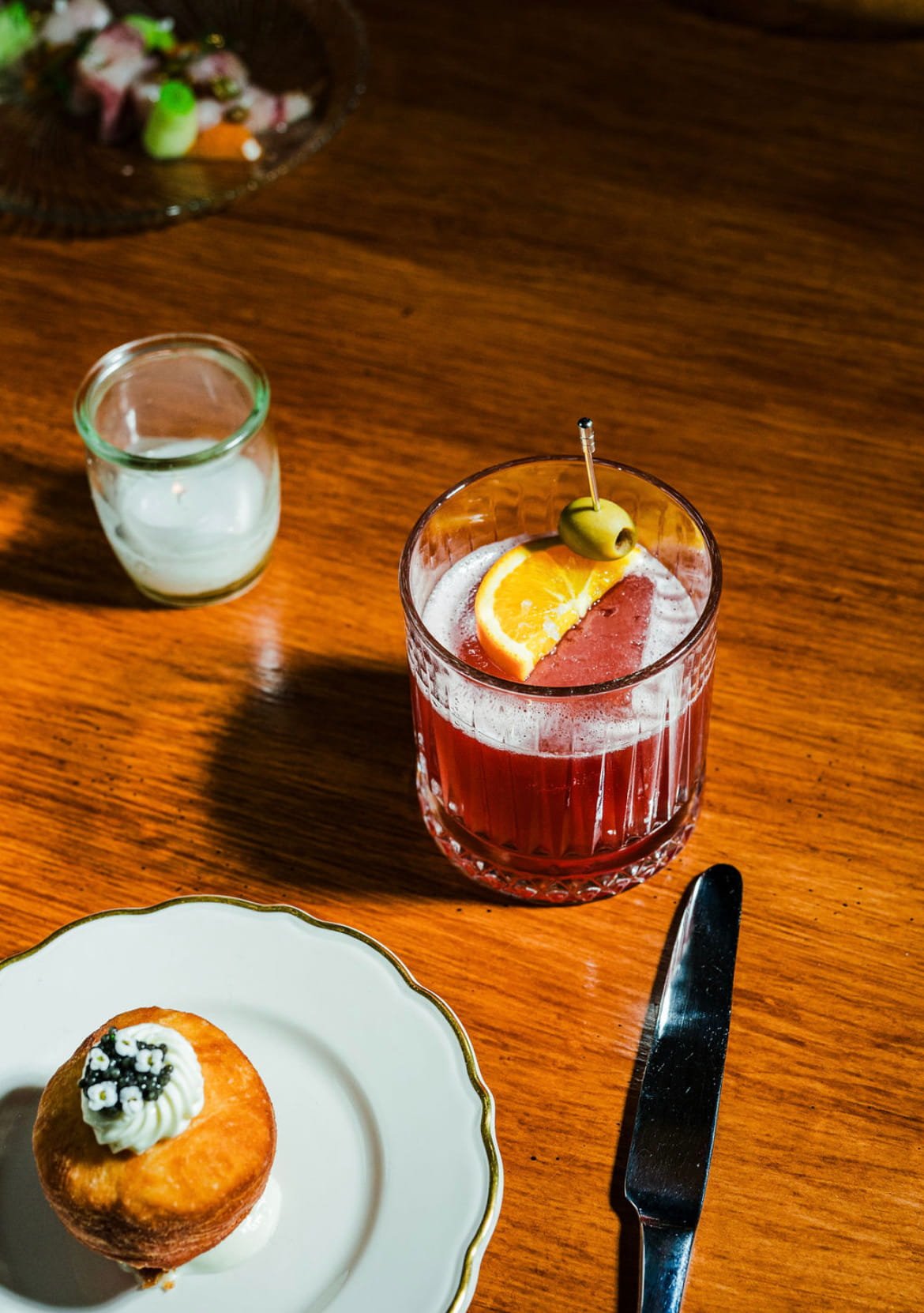 A glass of red cocktail with orange slice, green olive, and cherry on a wooden table, a small jar of cream, a cupcake with white frosting and black and white sprinkles, a knife, and part of a white plate with a gold rim in the scene.
