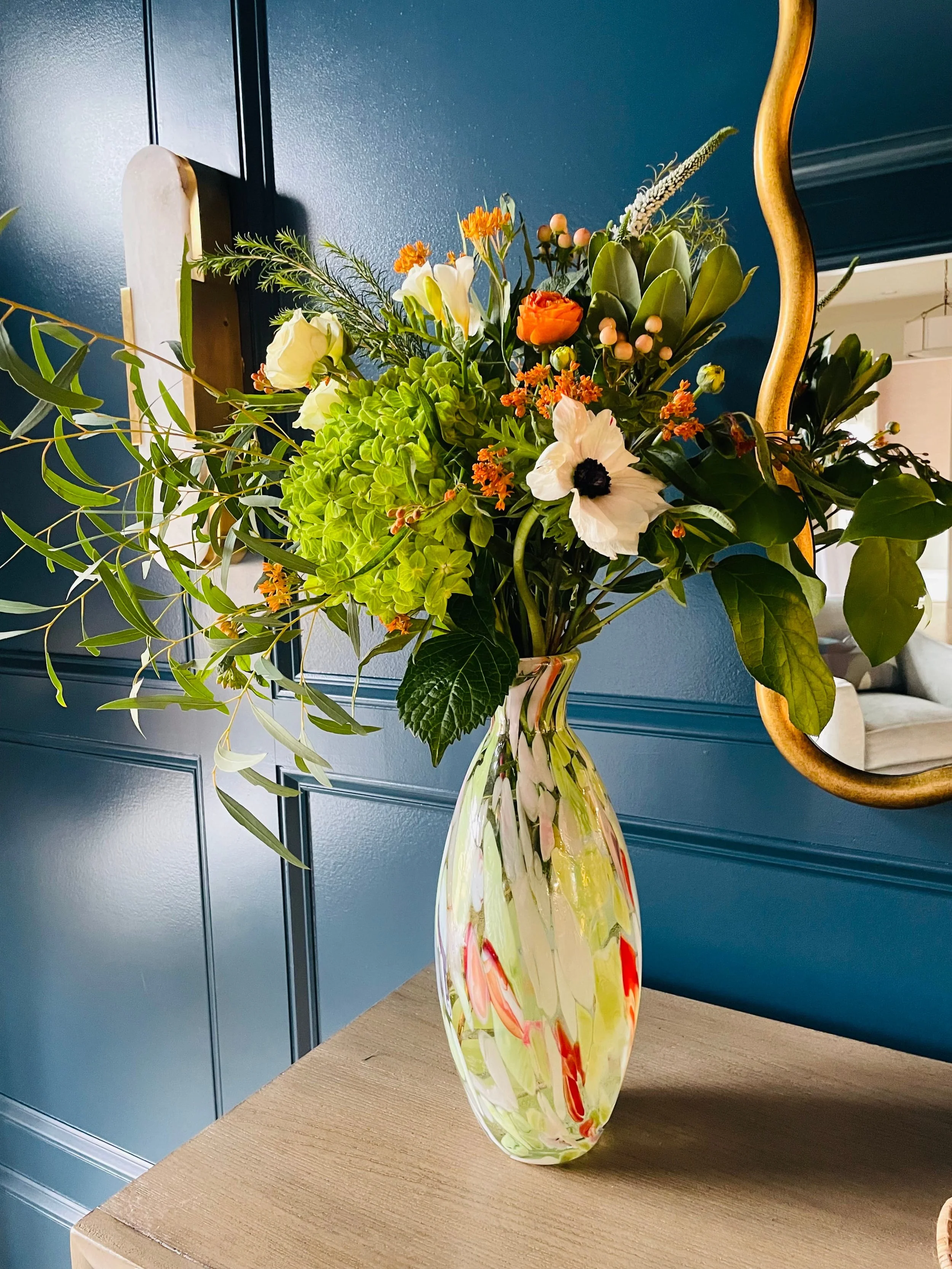 A tall, multicolored glass vase filled with a large arrangement of various flowers and greenery, placed on a wooden surface against a blue paneled wall.