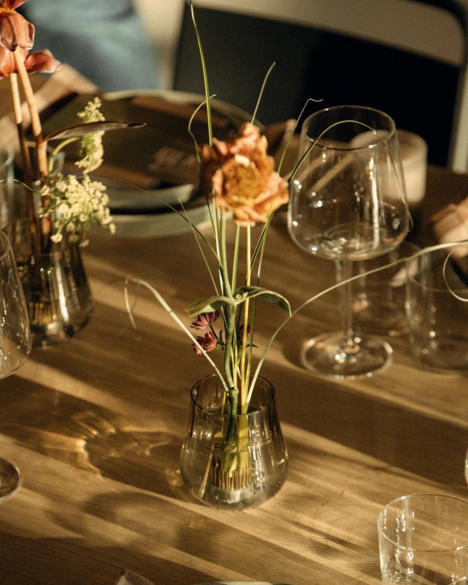 A small glass vase with a few flowers, including a pinkish flower and some green grass-like stems, sits on a wooden table. The table also has various empty wine glasses and a notepad or menu in the background.