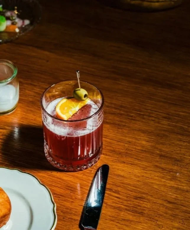 A glass of red cocktail with an orange slice and green olive garnish on a wooden table, with a butter knife and a plate partially visible.