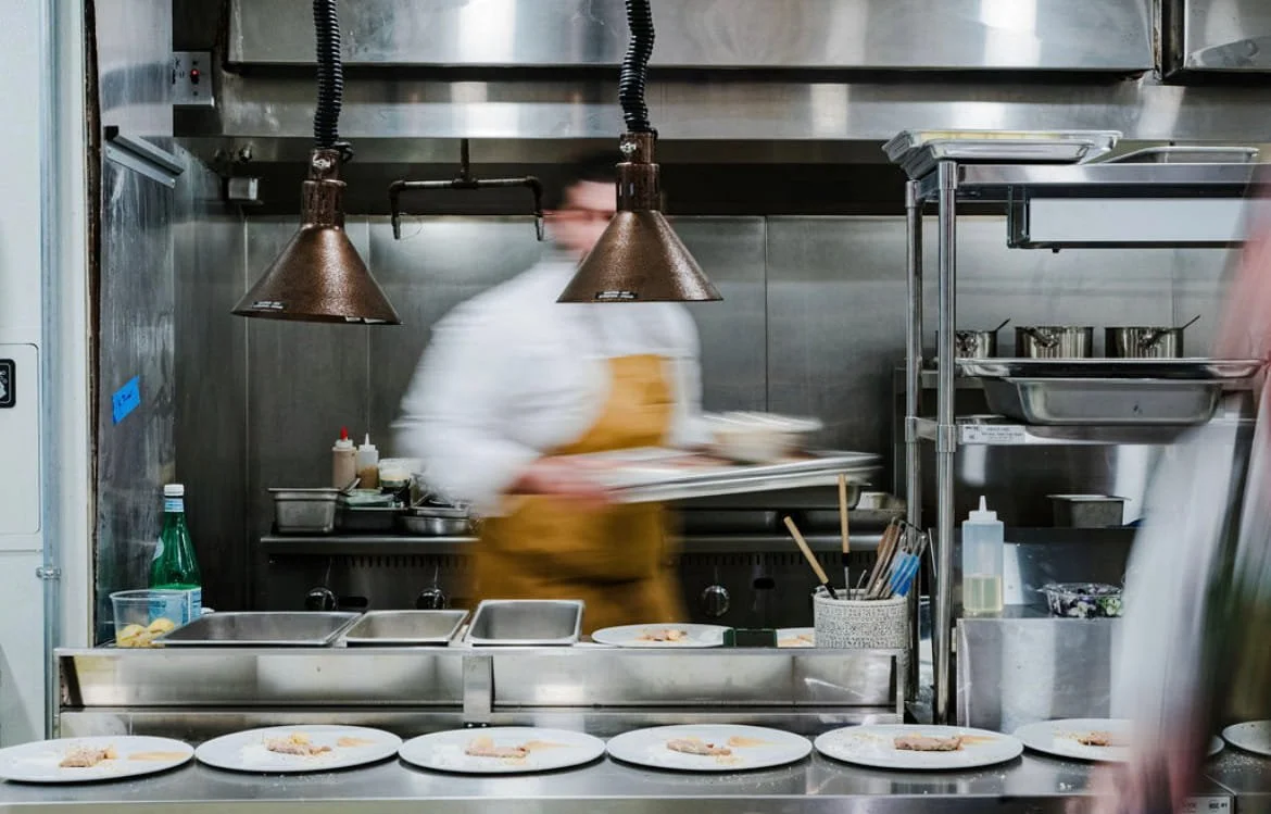 Blurry chef in a kitchen preparing plates of food with grills, utensils, and ingredients visible.