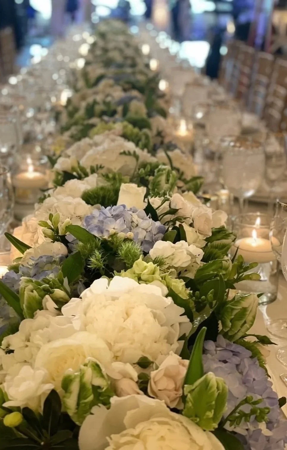 A close-up of a long floral centerpiece on a table, featuring white, pale yellow, and light purple flowers, illuminated by candlelight, with a blurred background.