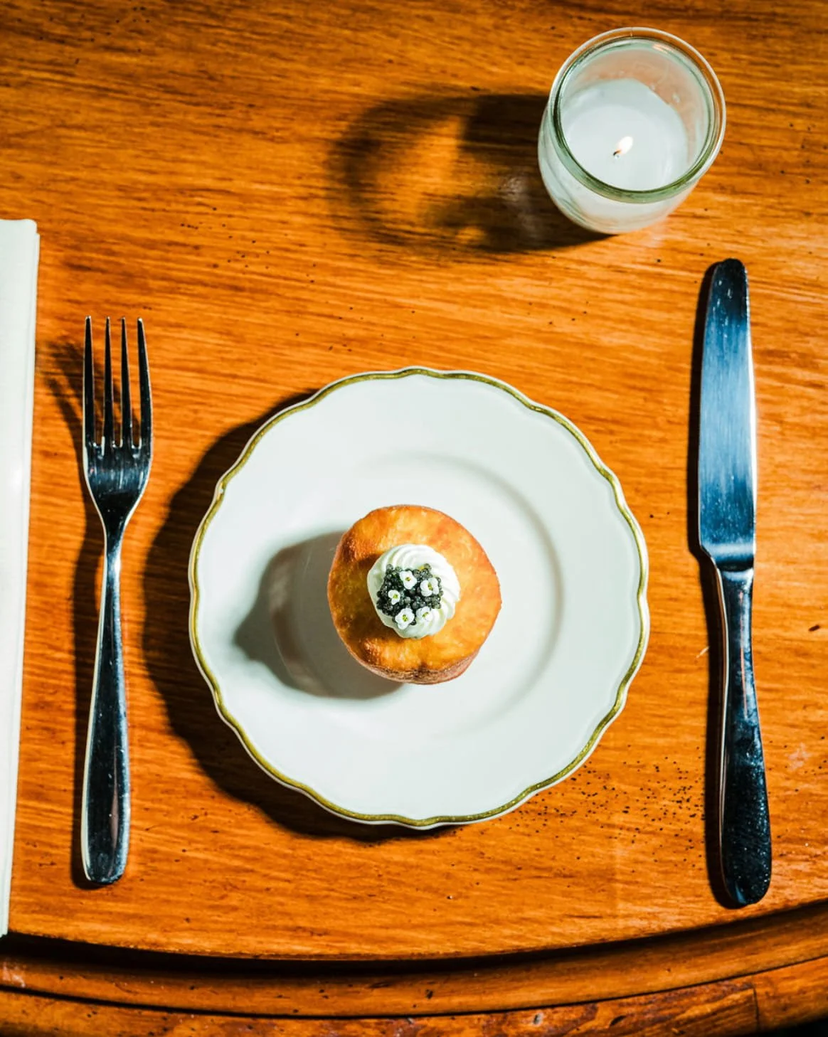 A small pastry with a dollop of cream and small flowers on top, placed on a white decorative plate, with a fork on the left and a knife on the right, a candle above on a wooden table.