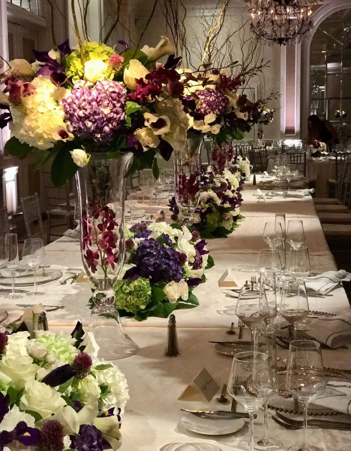 Elegant dining table decorated with a large floral centerpiece in a tall glass vase, featuring purple, pink, white, and green flowers, set for a formal event with wine glasses, silverware, napkins, and place cards in a decorated banquet hall.
