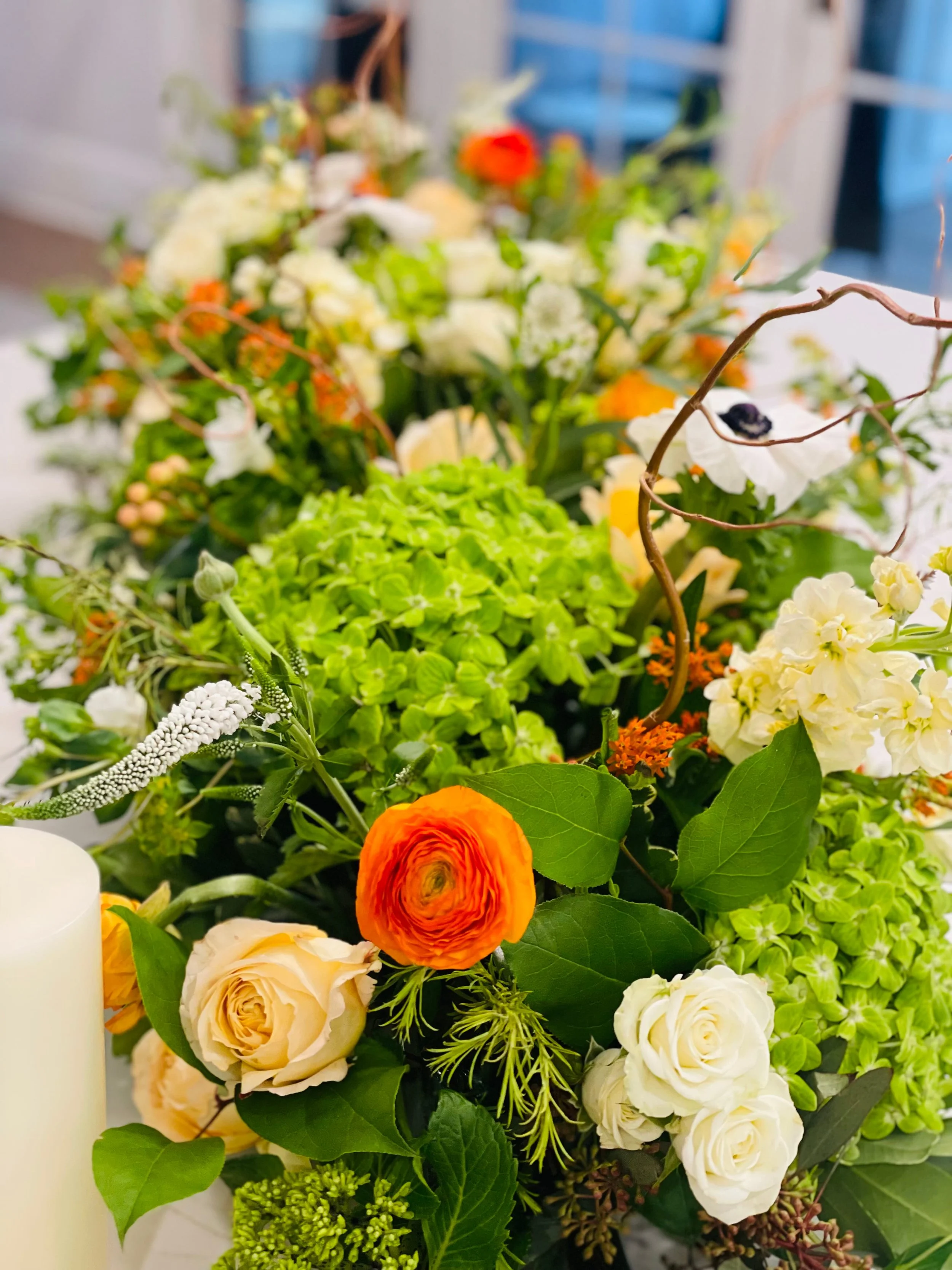 A floral arrangement featuring white, orange, and cream-colored flowers, along with green foliage and some curly birch branches, with blurred background.