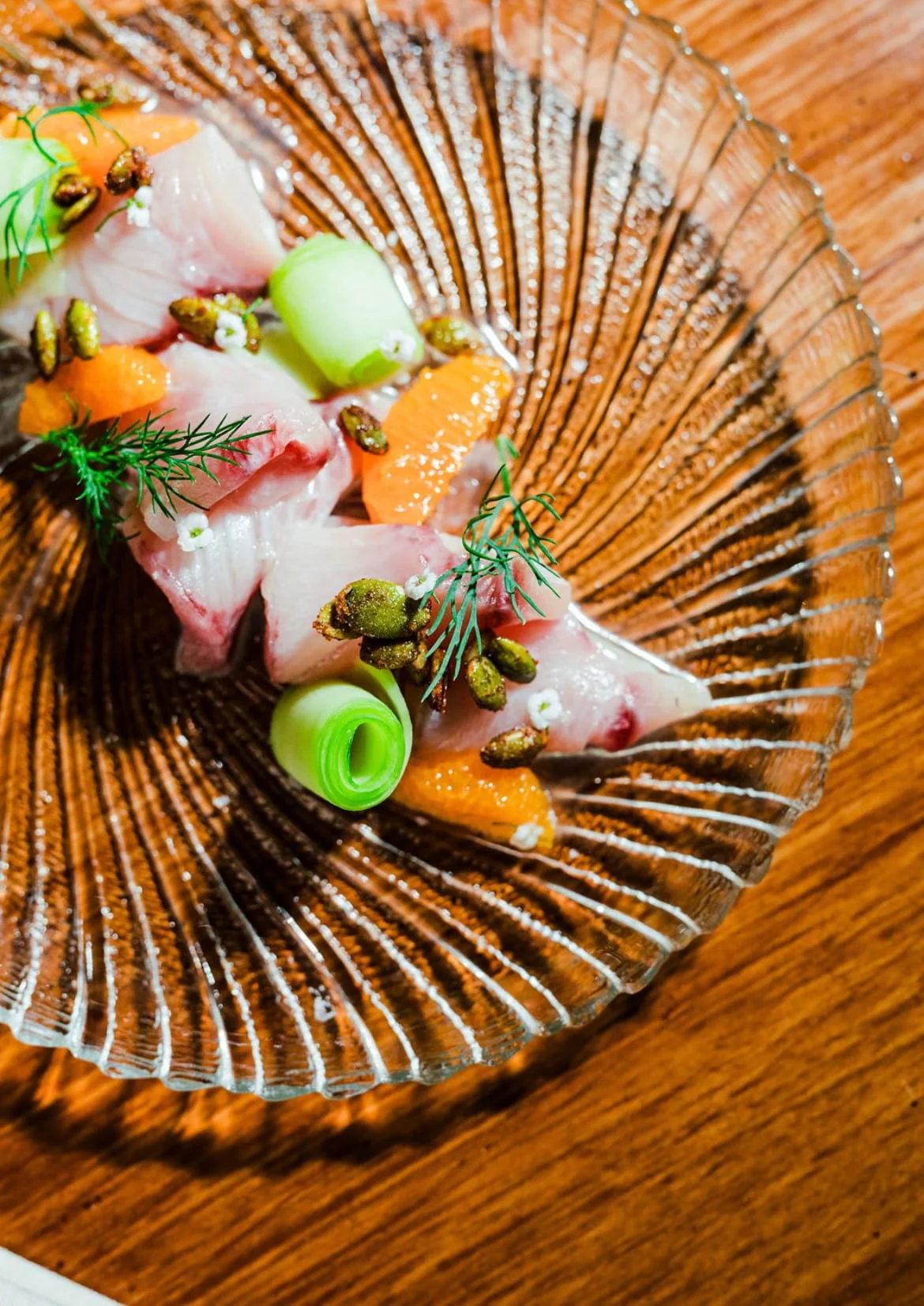 Close-up of a glass dish with raw fish, orange carrot slices, green scallion, garnished with herbs, small flowers, and edible seeds on a wooden surface.