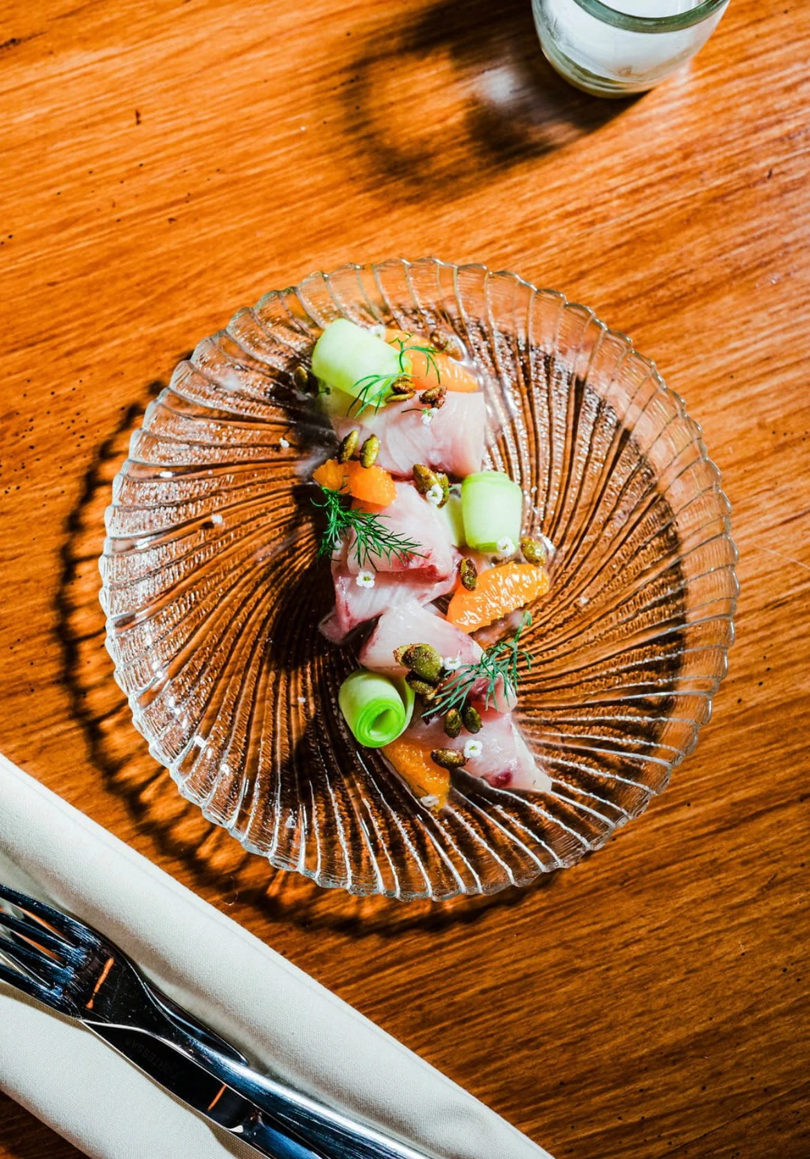 Sashimi platter with slices of raw fish, cucumber rolls, small edible flowers, herbs, and garnishes on a glass plate.
