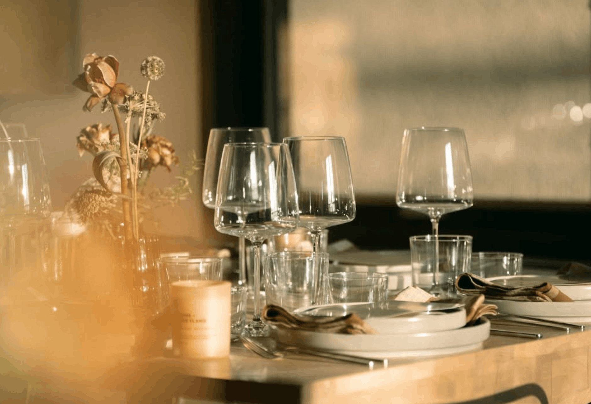 A dining table is set with empty wine glasses, plates, napkins, and cutlery, with a centerpiece of dried flowers in a vase and warm lighting creating a cozy ambiance.