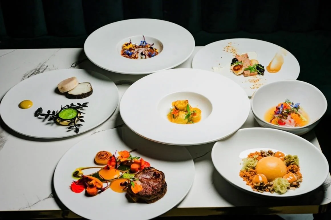 Assorted gourmet dishes on white plates arranged on a marble table, featuring various colorful and intricately presented appetizers and entrees.