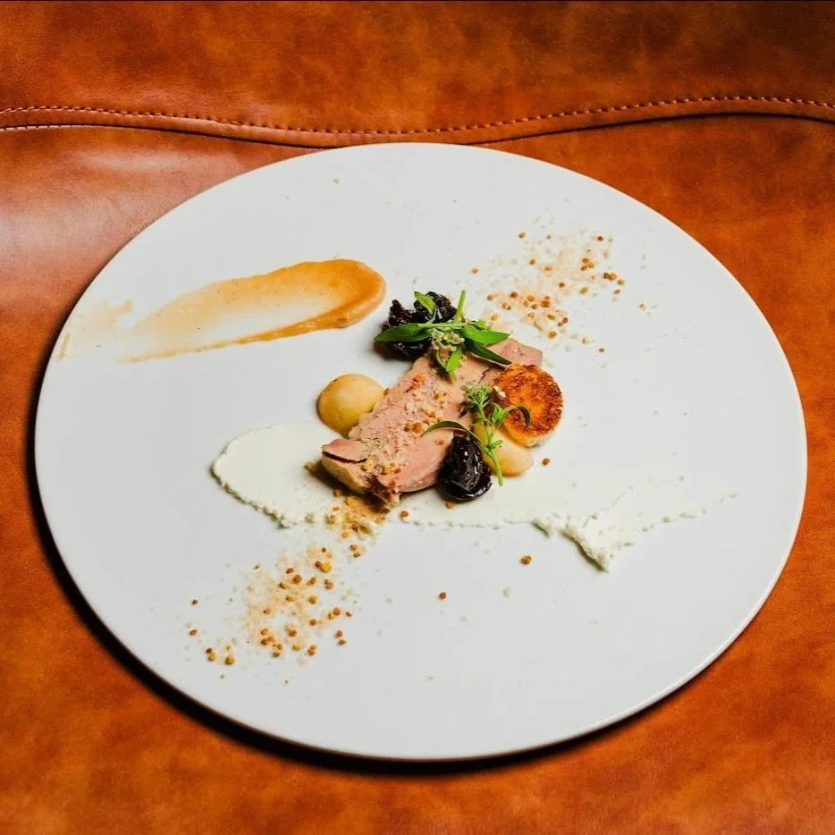 A white plate with a small serving of sliced meat, garnished with greens, a dollop of white sauce, a smear of brown sauce, and sprinkled spices, on a wooden surface.