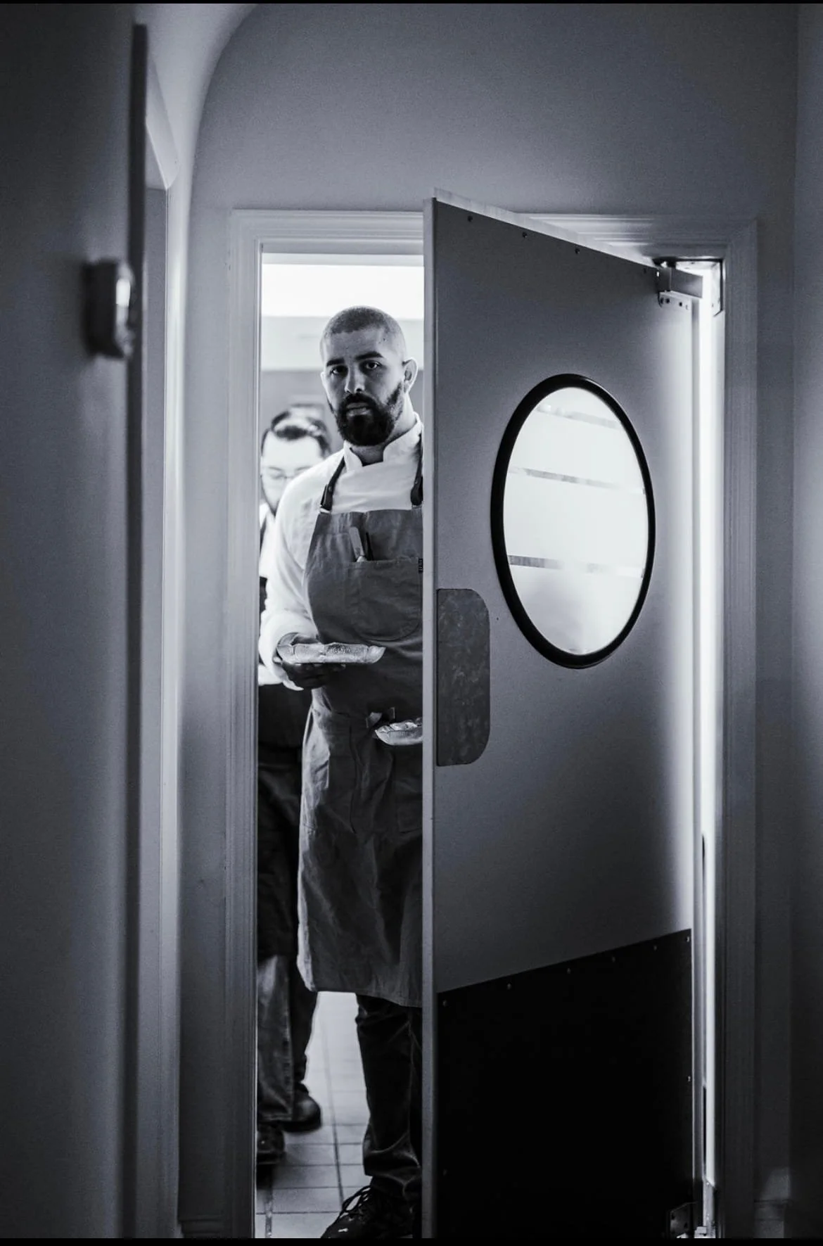 A chef with a beard and short hair, wearing an apron, stands in a doorway holding a plate. Behind him is another person in the kitchen. The doorway has a round window and is partially open, leading into a brightly lit area.