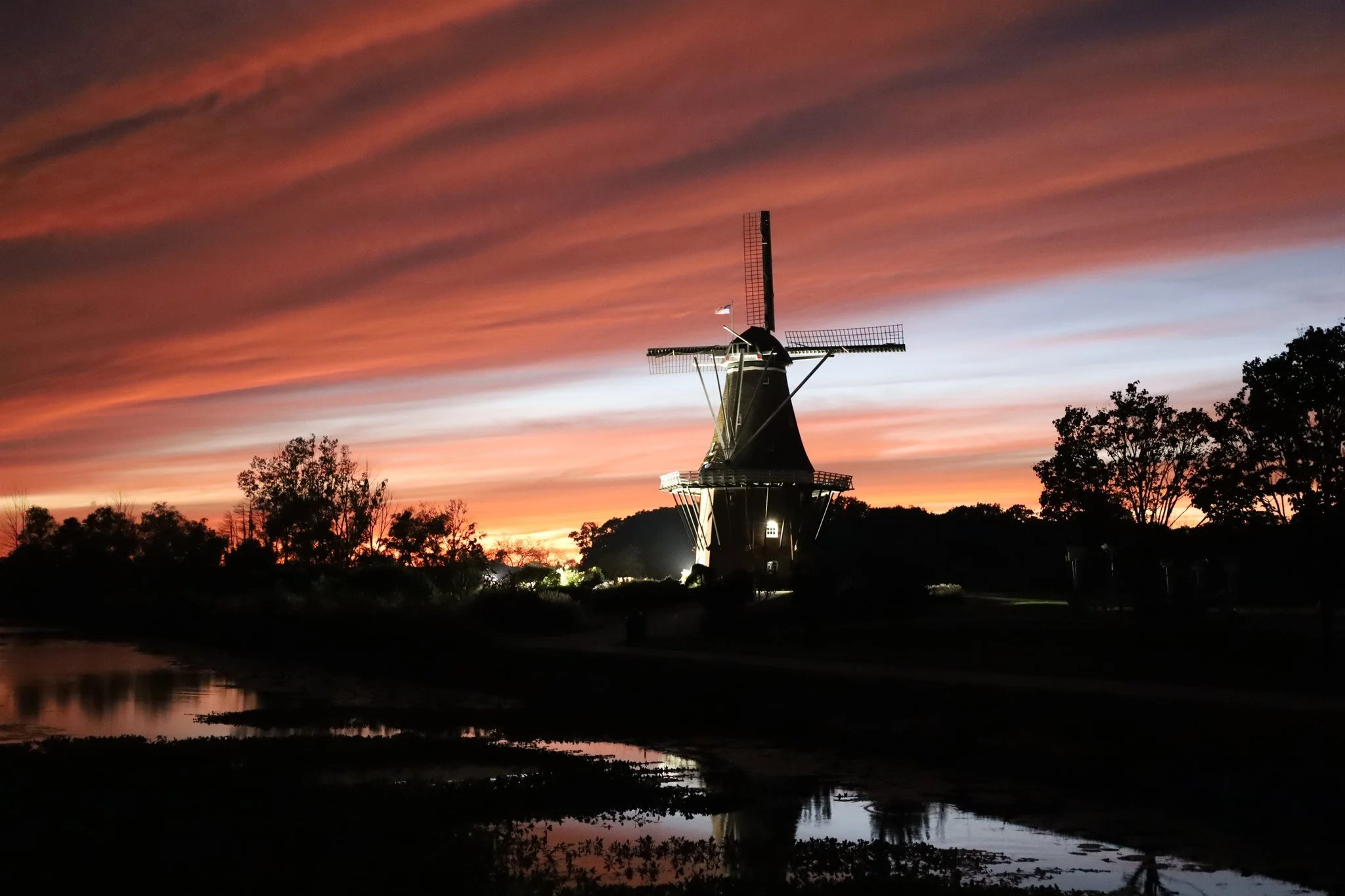 Silhouette of a traditional Dutch windmill against a colorful sunset sky with clouds and trees in the background.