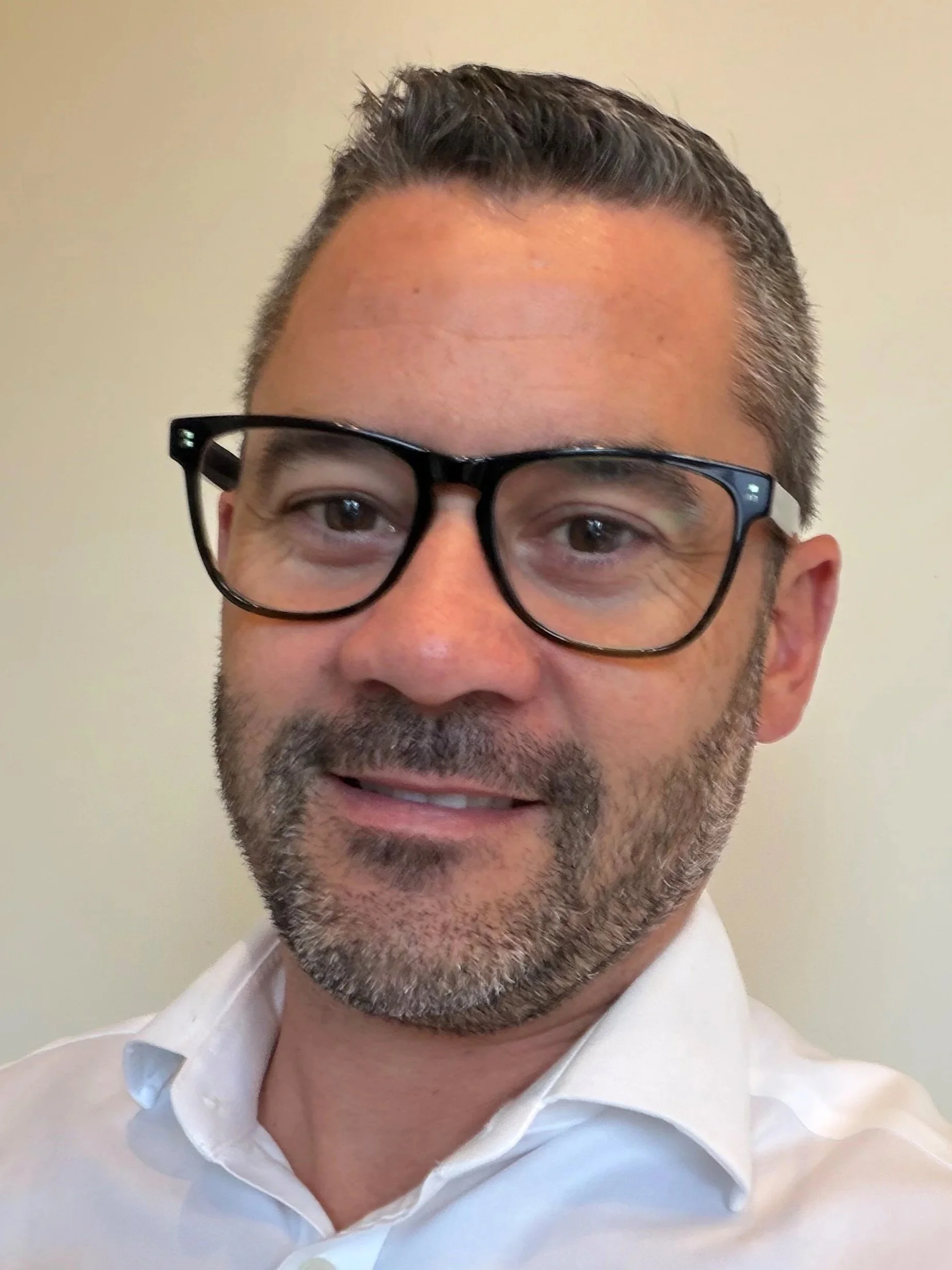 A man with short dark hair, a beard, wearing black-rimmed glasses and a white collared shirt, smiling gently at the camera against a plain light-colored background.