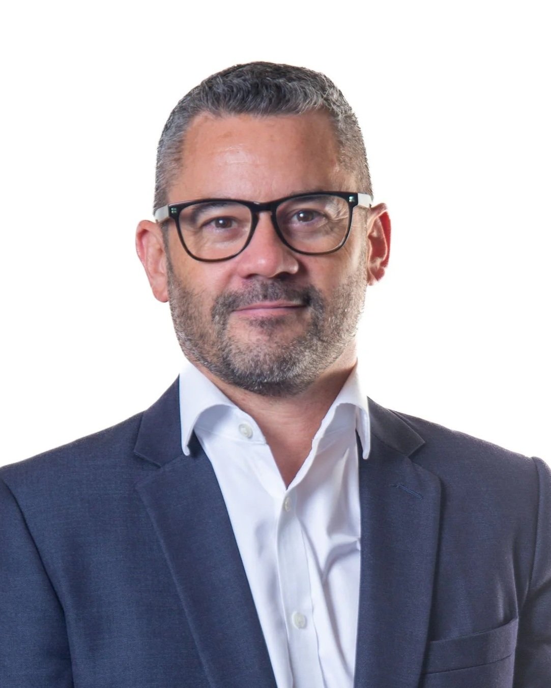 A man with short gray hair, wearing black-rimmed glasses, a white shirt, and a navy blazer, posing against a plain white background.