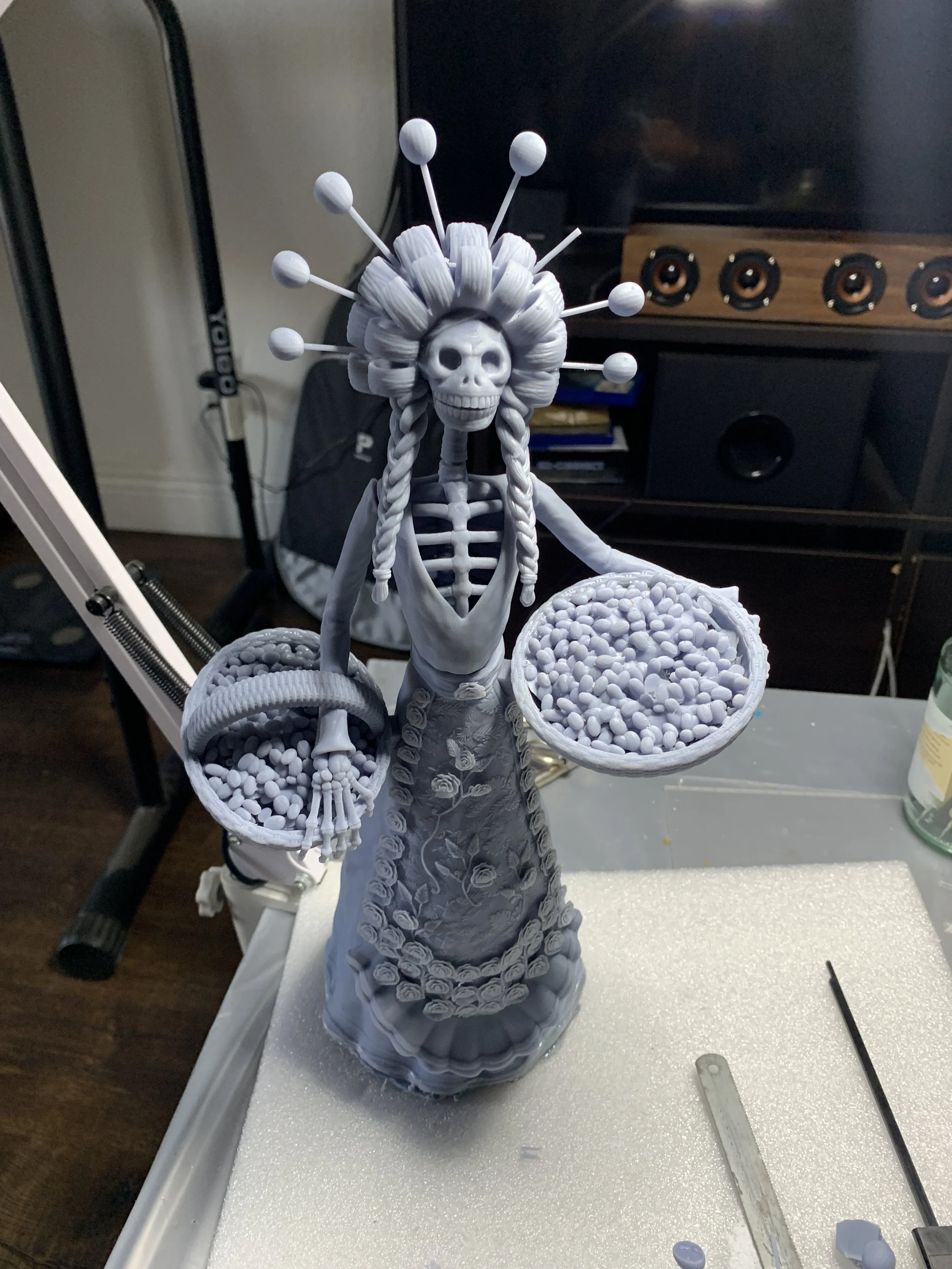 A sculpted figure featuring a skeletal woman with intricate hair decoration and braided hair, holding two baskets filled with small items. The figure is adorned in a detailed dress with floral patterns. The sculpture is in a workshop setting, with to