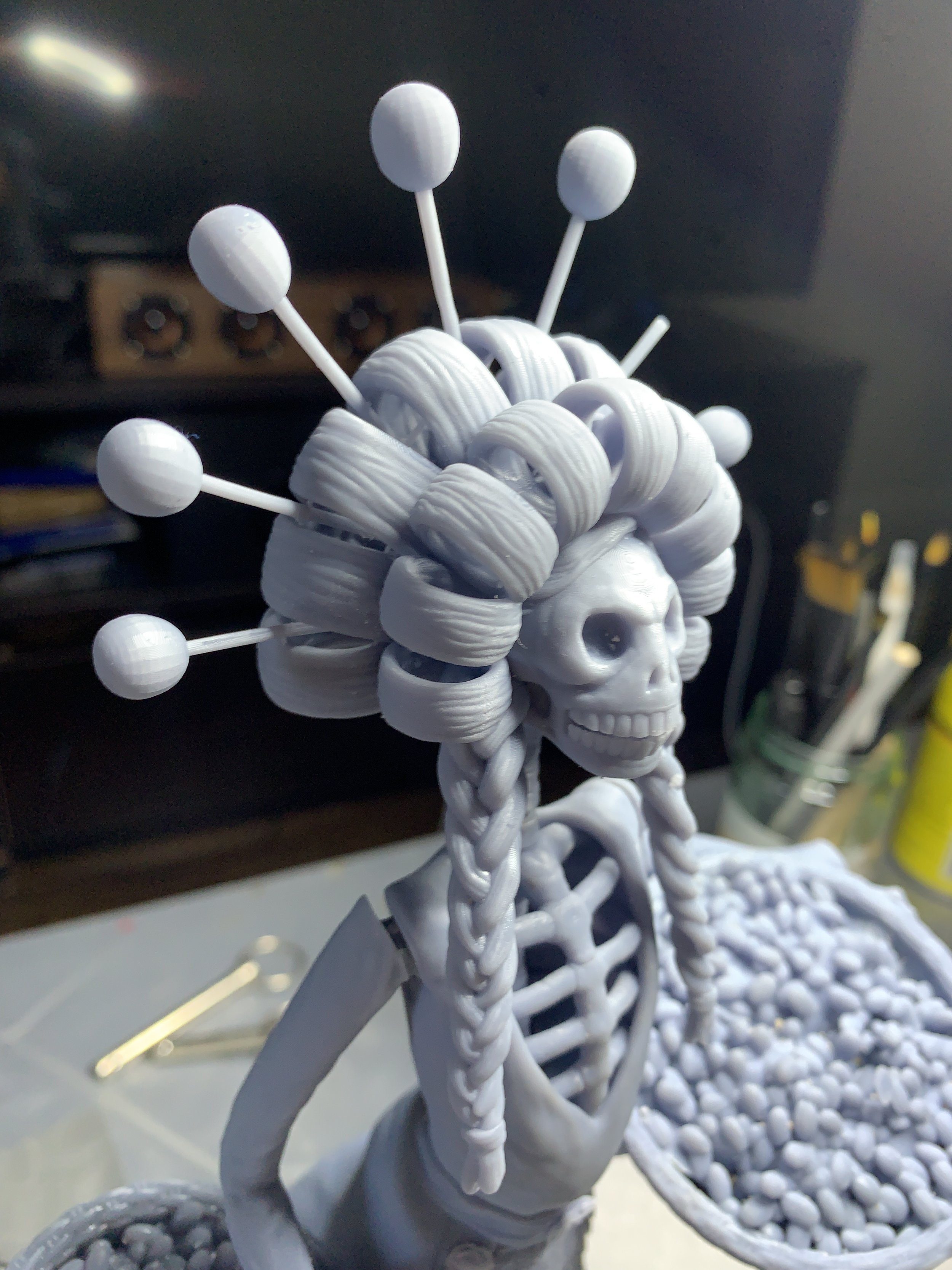 3D printed skeletal figure wearing a braided wig with round ornaments, resembling a stylized character or sculpture. The figure is in a workshop setting with tools and materials in the background.