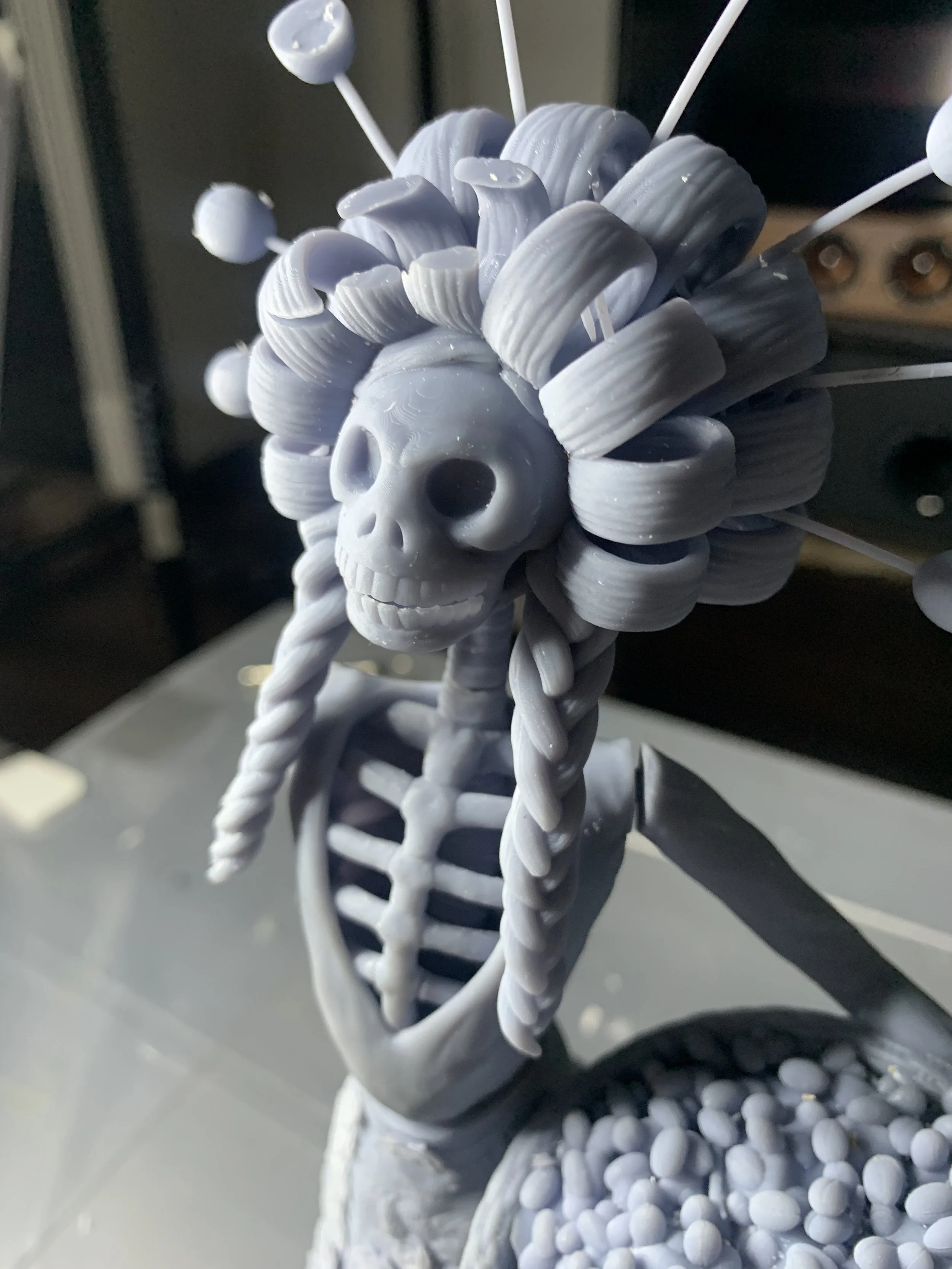 A close-up of a sculpted figure featuring a skeletal face with hollow eyes and a toothy grin. The figure has a complex headdress made of coiled and looped structures, resembling hair. It includes stylized braids and decorative elements protruding fro