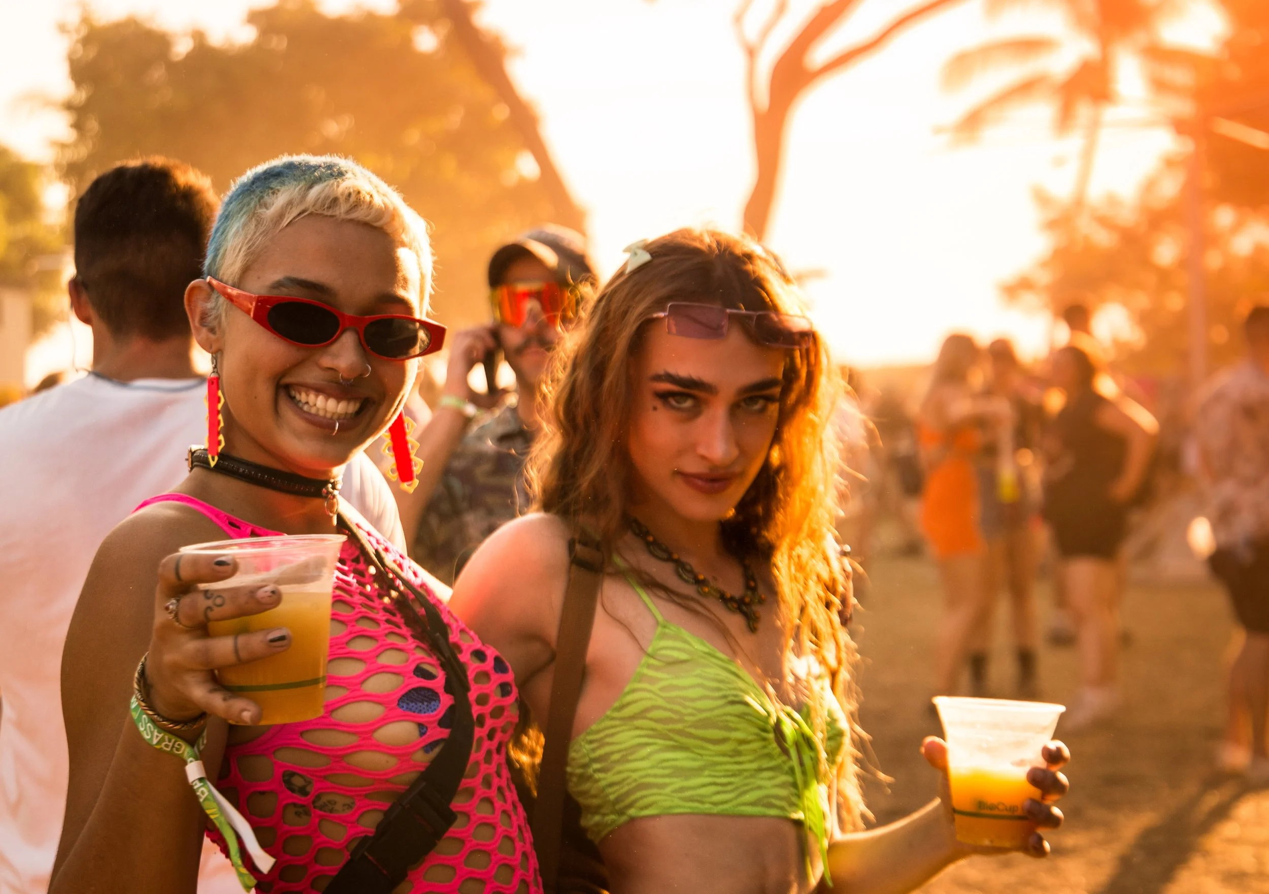 Two women at an outdoor event during sunset, both holding drinks and smiling, with others in the background.  They are at the Bass in the Grass Festival in Darwin in the Northern Territory.