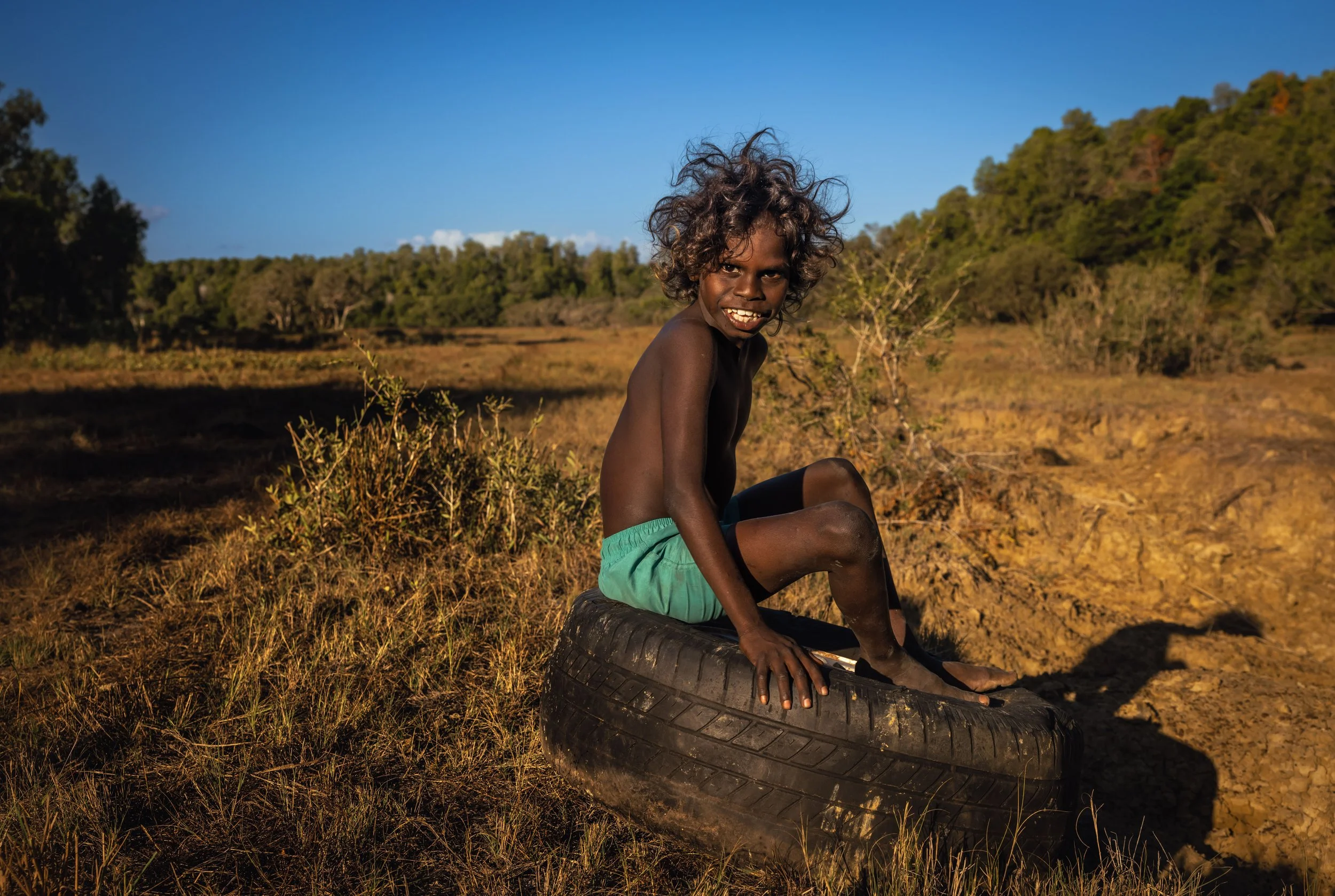A young boy with curly hair, no shirt, green shorts, sitting on a used tire in a dry grassy field with trees in the distance under a clear blue sky. An Arnhem Land portrait of life in the NT.