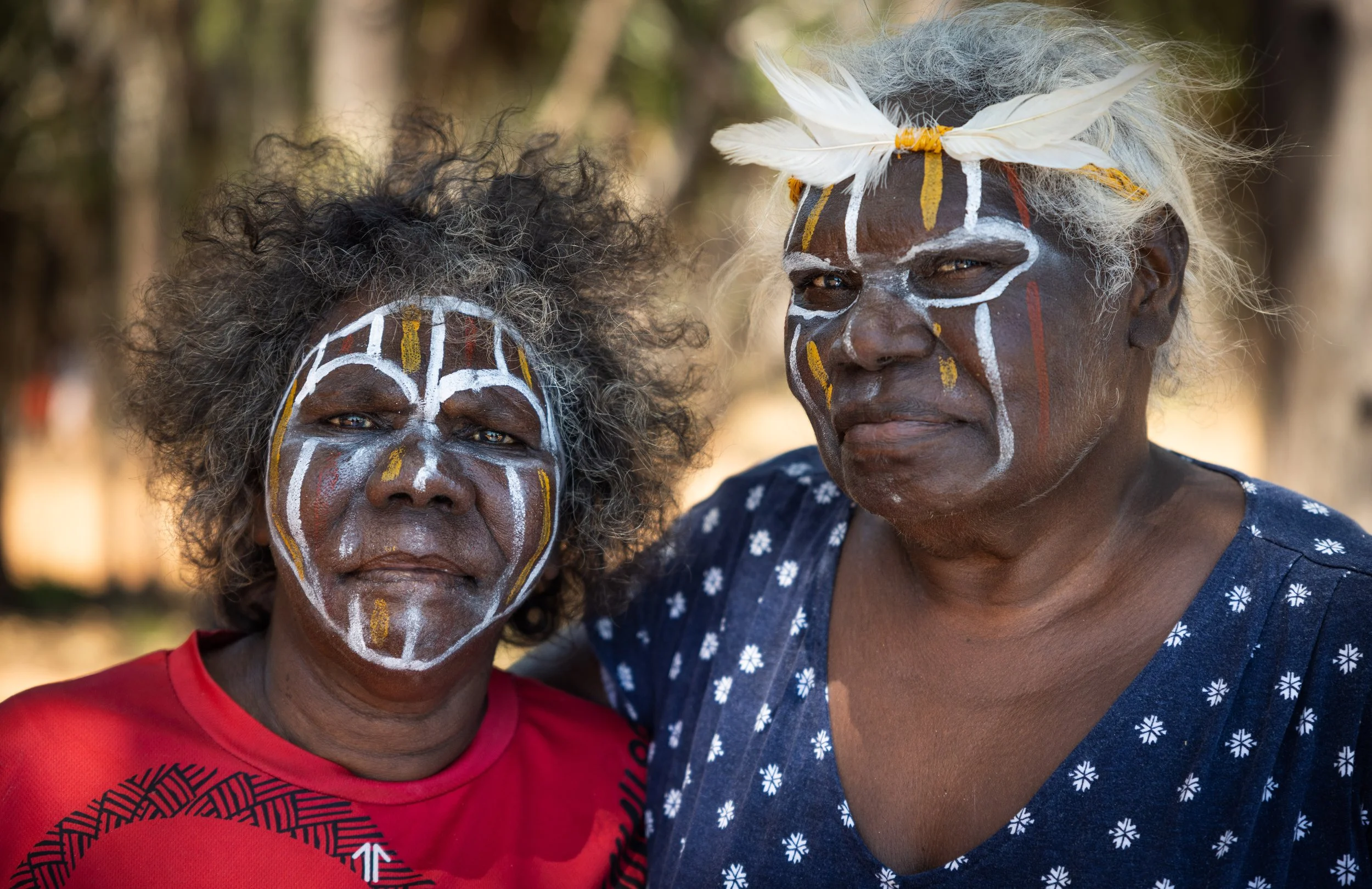 Two Tiwi women with traditional face paint, one wearing a red shirt and the other a blue shirt and a headband with feathers.