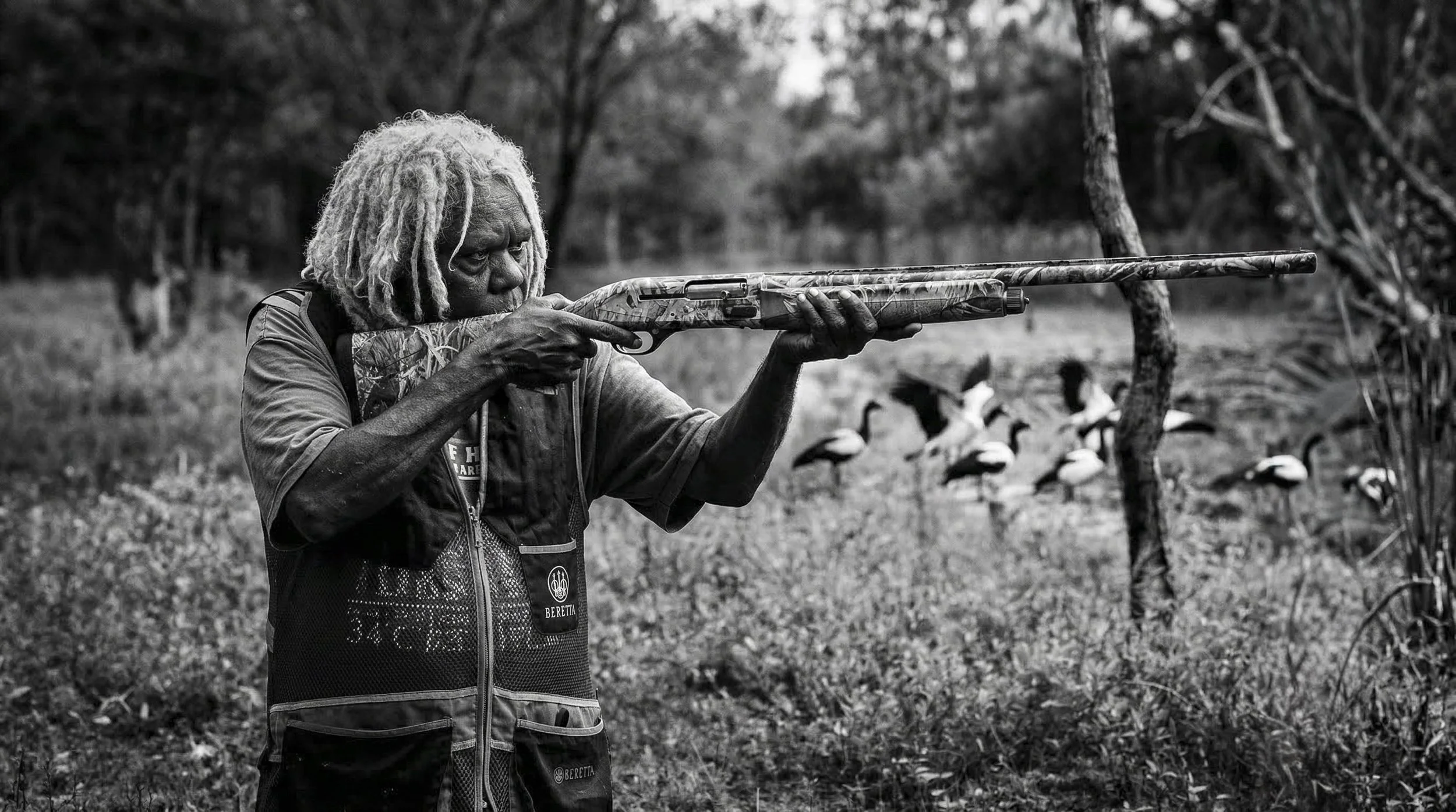 An elderly First Nations woman with dreadlocks aiming a camouflage rifle outdoors, surrounded by trees and Lake in background, with Magpie Geese on the swamp.  A portrait from Kakadu in the Northern Territory.