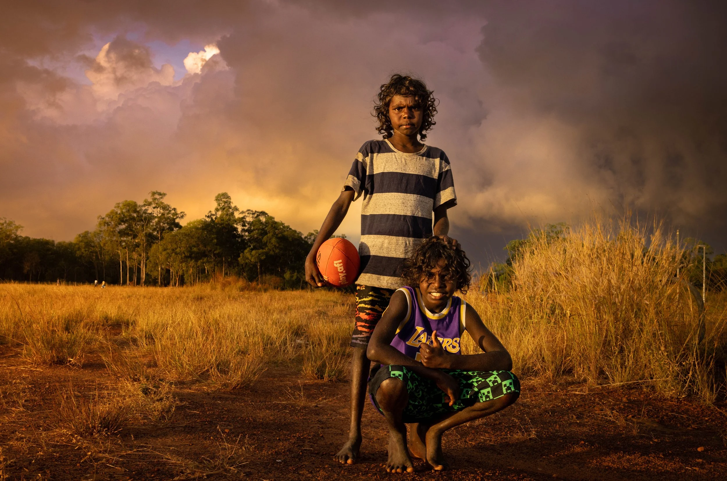 Two young First Nations boys in casual clothes in an open field with tall grass and trees, one holding a basketball, under a dramatic cloudy sky during sunset.