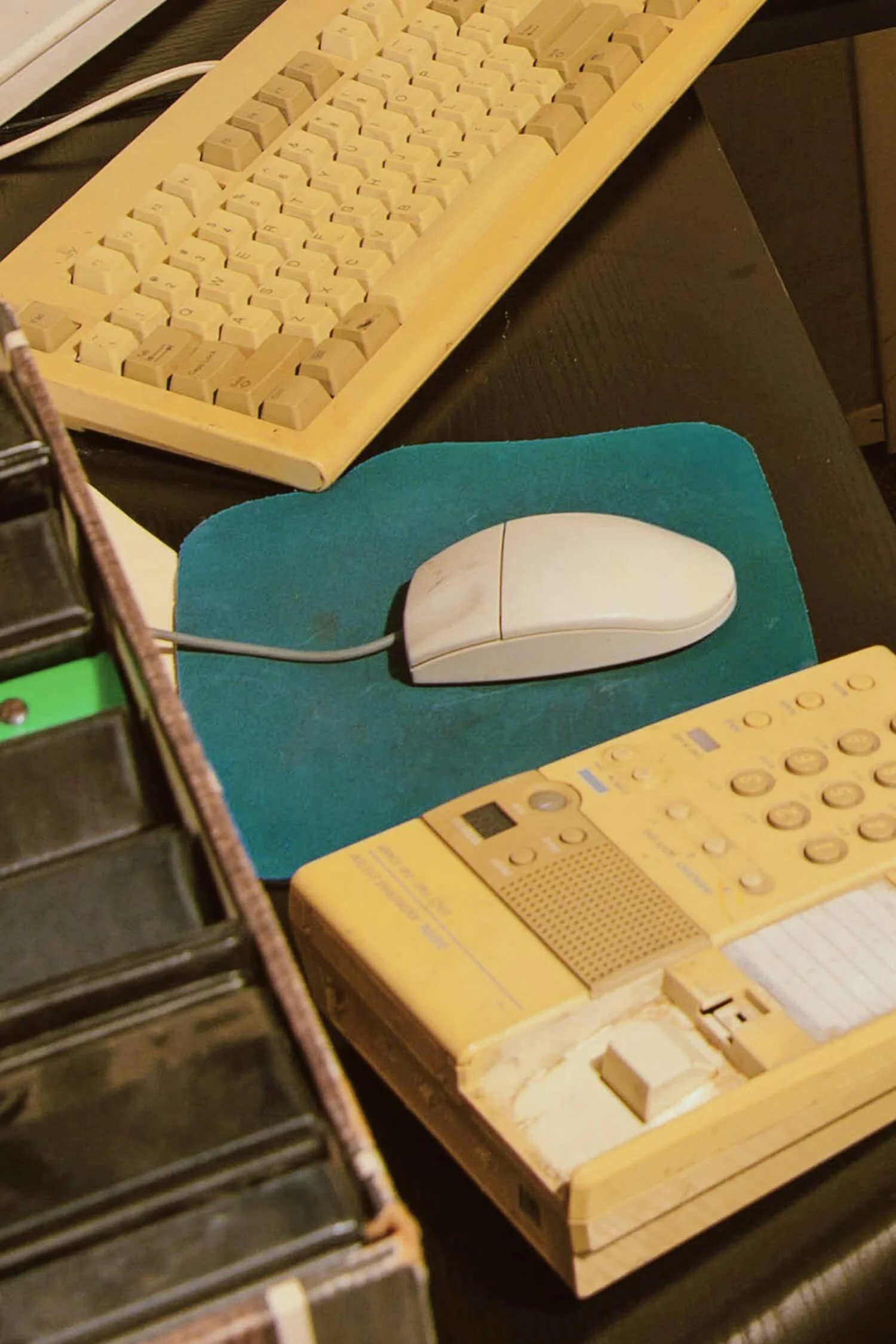 An old computer keyboard, a computer mouse on a green mouse pad, and an electronic device with buttons and a small display on a dark desk.
