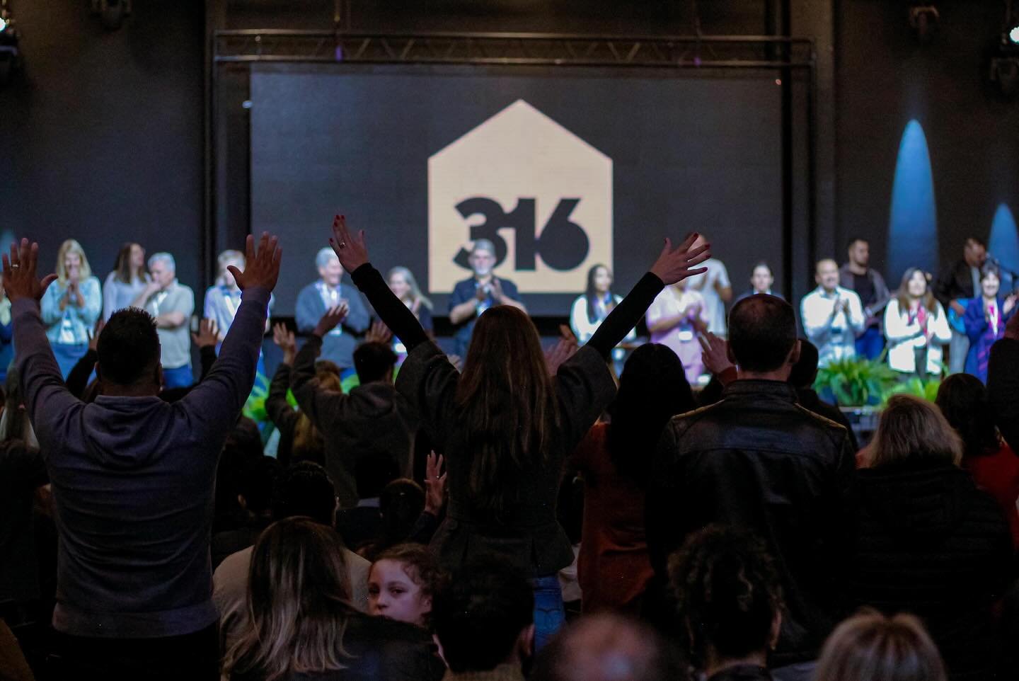 People on stage and in the audience at a conference or event, with a large screen showing '3K' in the background, some audience members with hands raised.