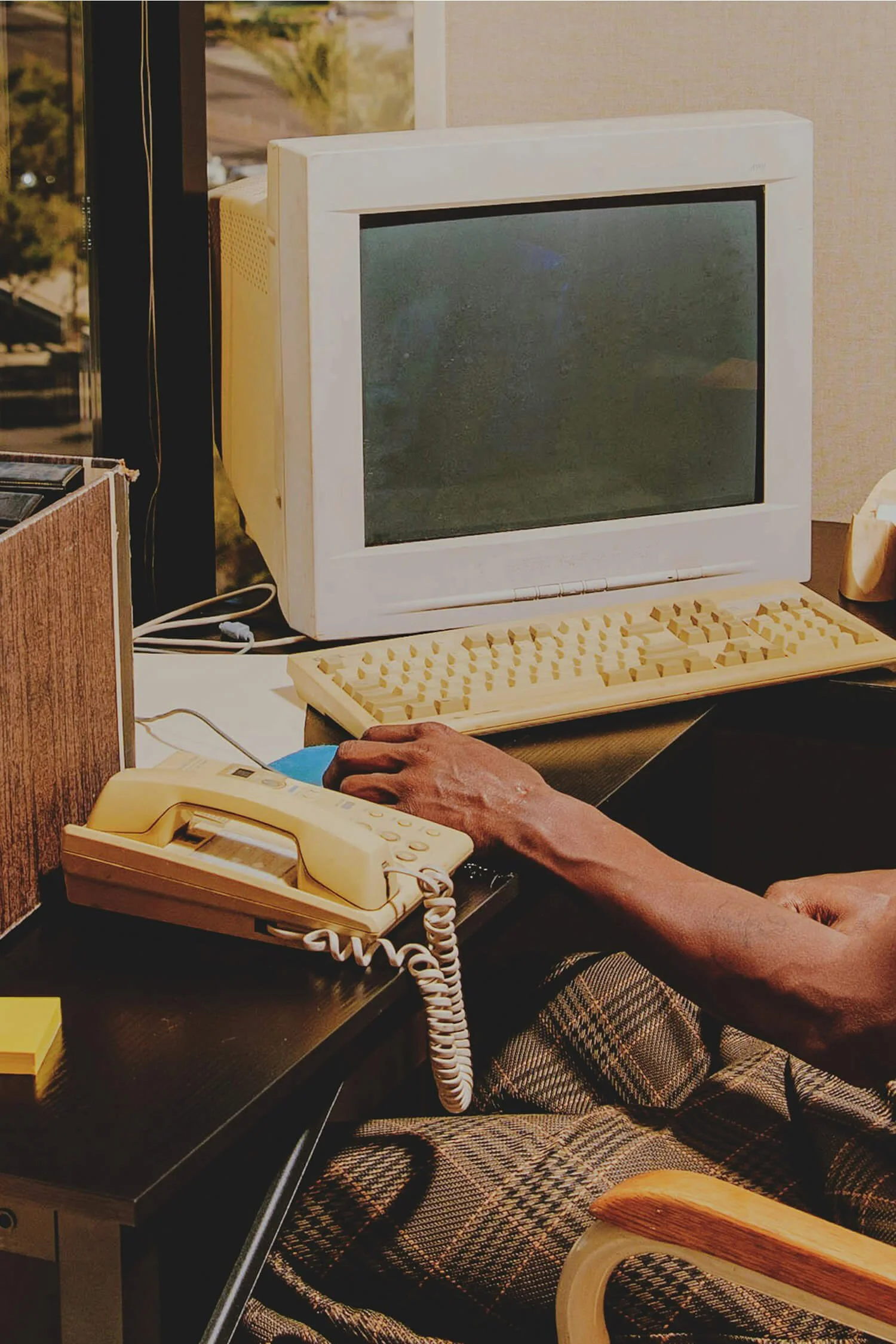 A vintage office desk with an old beige computer monitor, keyboard, and telephone, with a person's hand on the mouse, in a well-lit room.