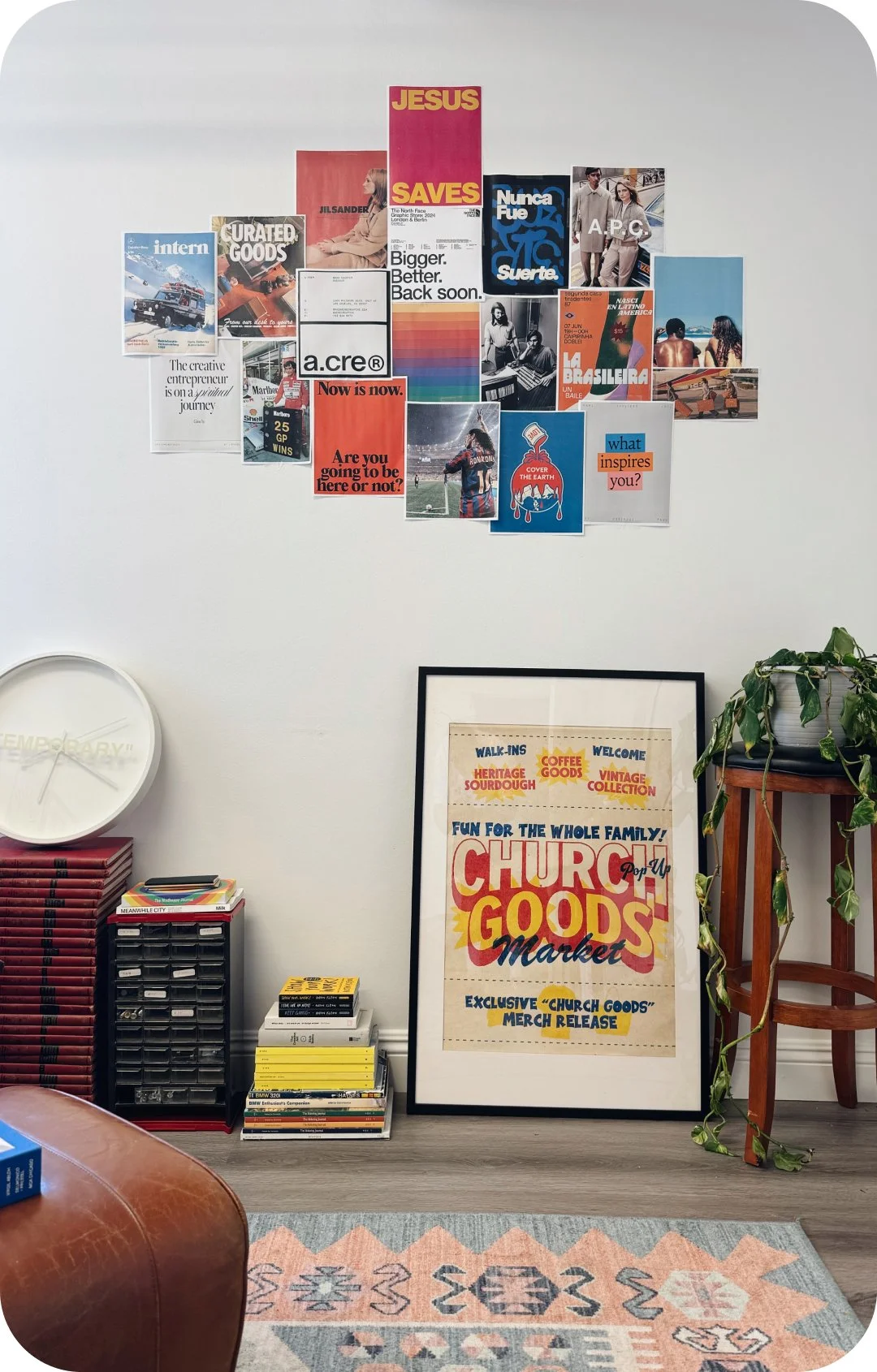 Room with a wall decorated with a collage of magazine clippings and posters, a framed sign reading 'Church Goods Market,' a plant on a wooden stand, stacks of magazines, and a round clock.