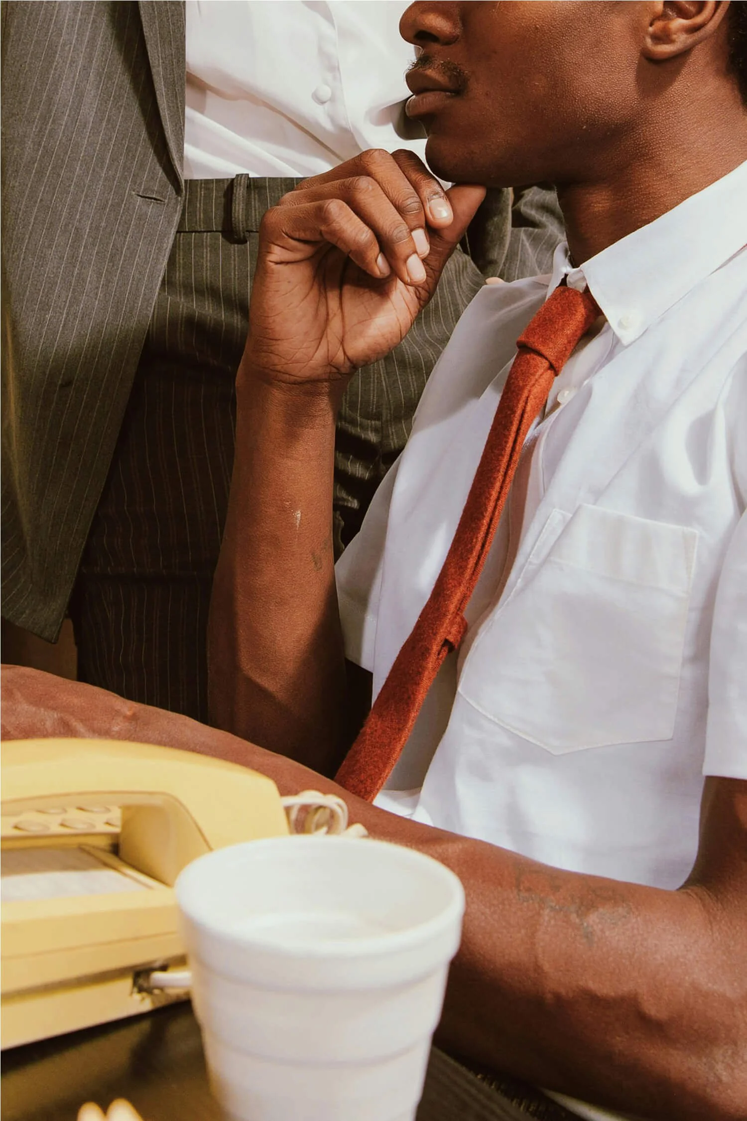 A man in a white shirt and tie, with a blurred person in a brown suit standing nearby, is shown with his chin resting on his hand, sitting at a desk with a yellow corded telephone and a white cup.