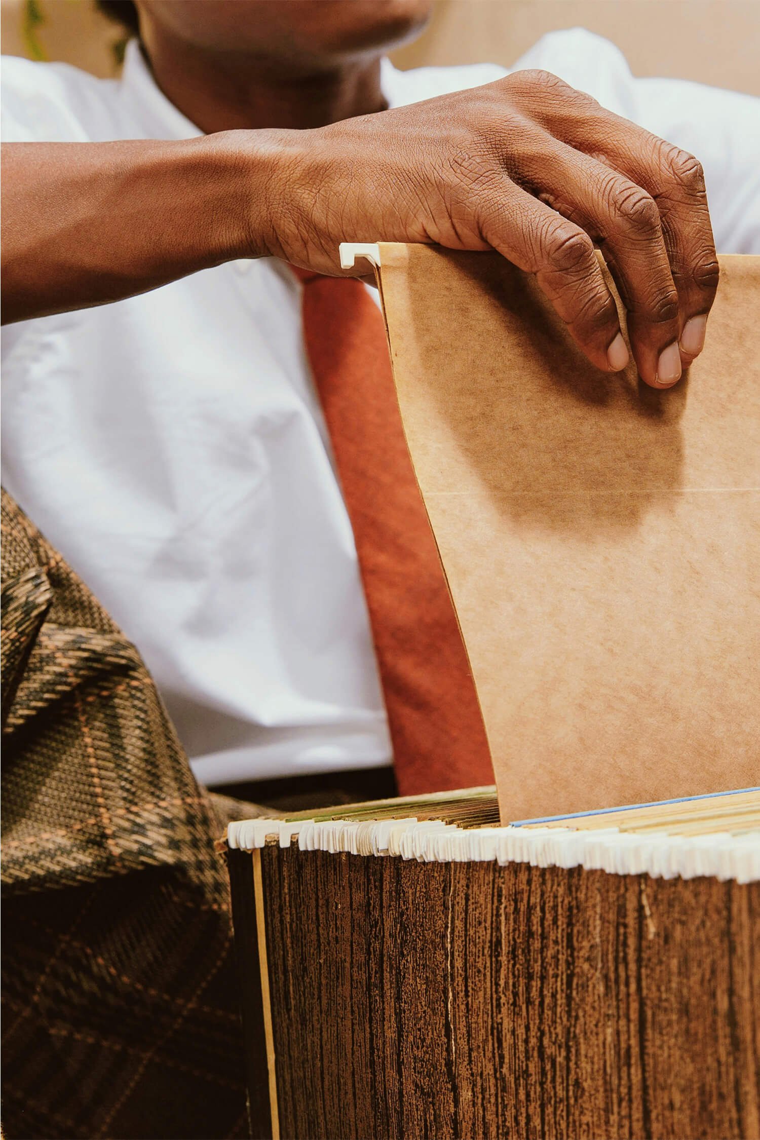 A person with dark skin is opening a brown paper bag over a table with a wooden box filled with coffee stirrers. The person is wearing a white shirt and an orange tie, and part of their face is visible.