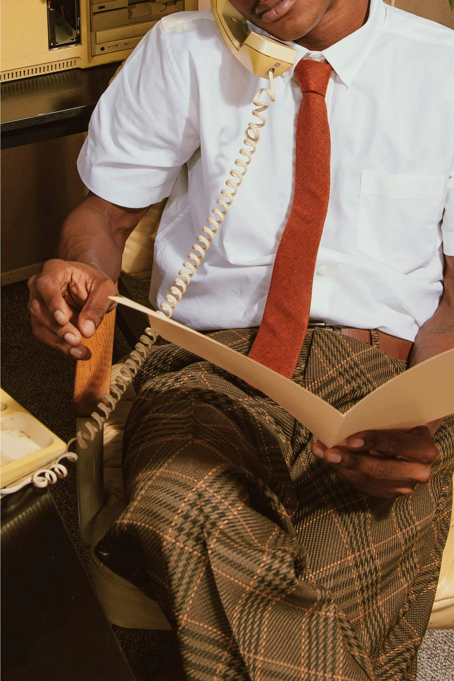 A man in a white shirt, patterned trousers, and a reddish-brown tie sitting at a desk, talking on a vintage beige corded telephone while reading a file or paper.