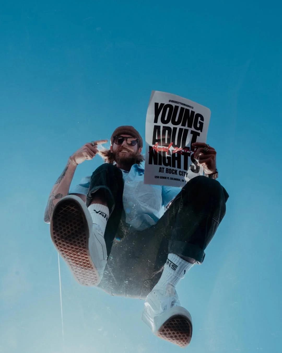 A man with a beard, sunglasses, and a beanie holds a sign that reads 'Young Adult Nights at Rock City' while sitting on skateboard against a blue sky.