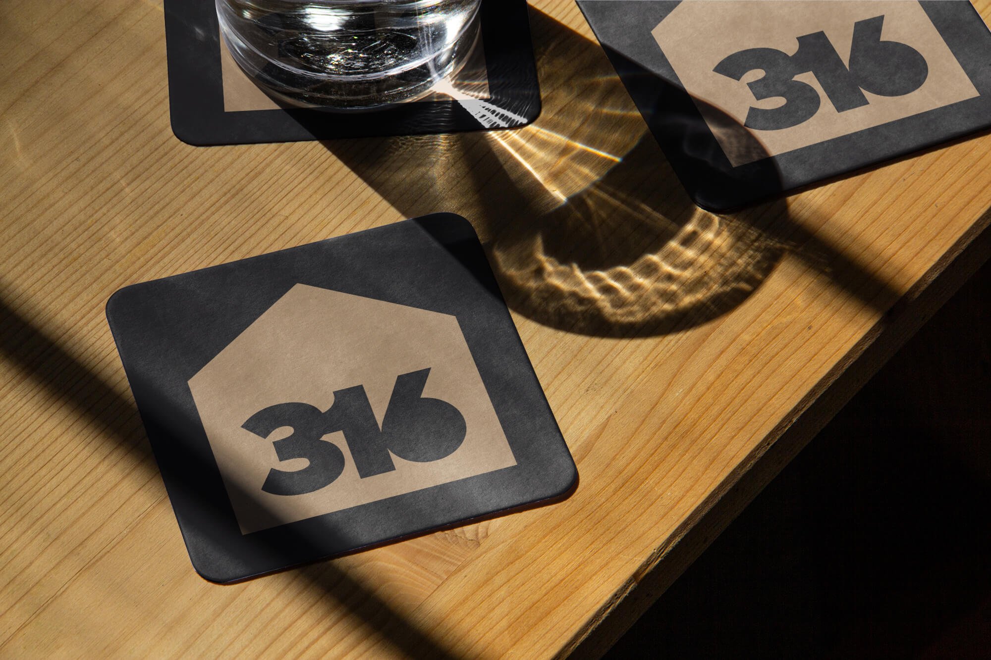 Coasters with a house logo and the text '3M' on a wooden table, with a glass of water and reflections from light.