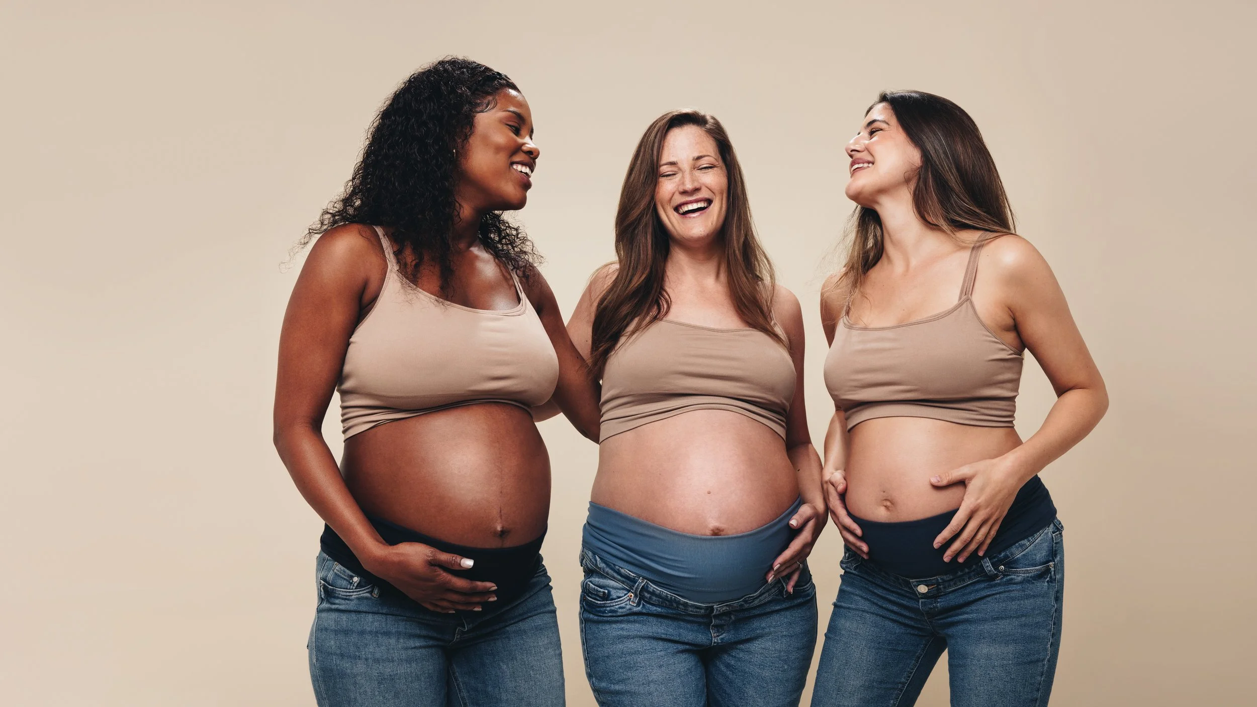 Three pregnant women standing together, each touching their belly and smiling.