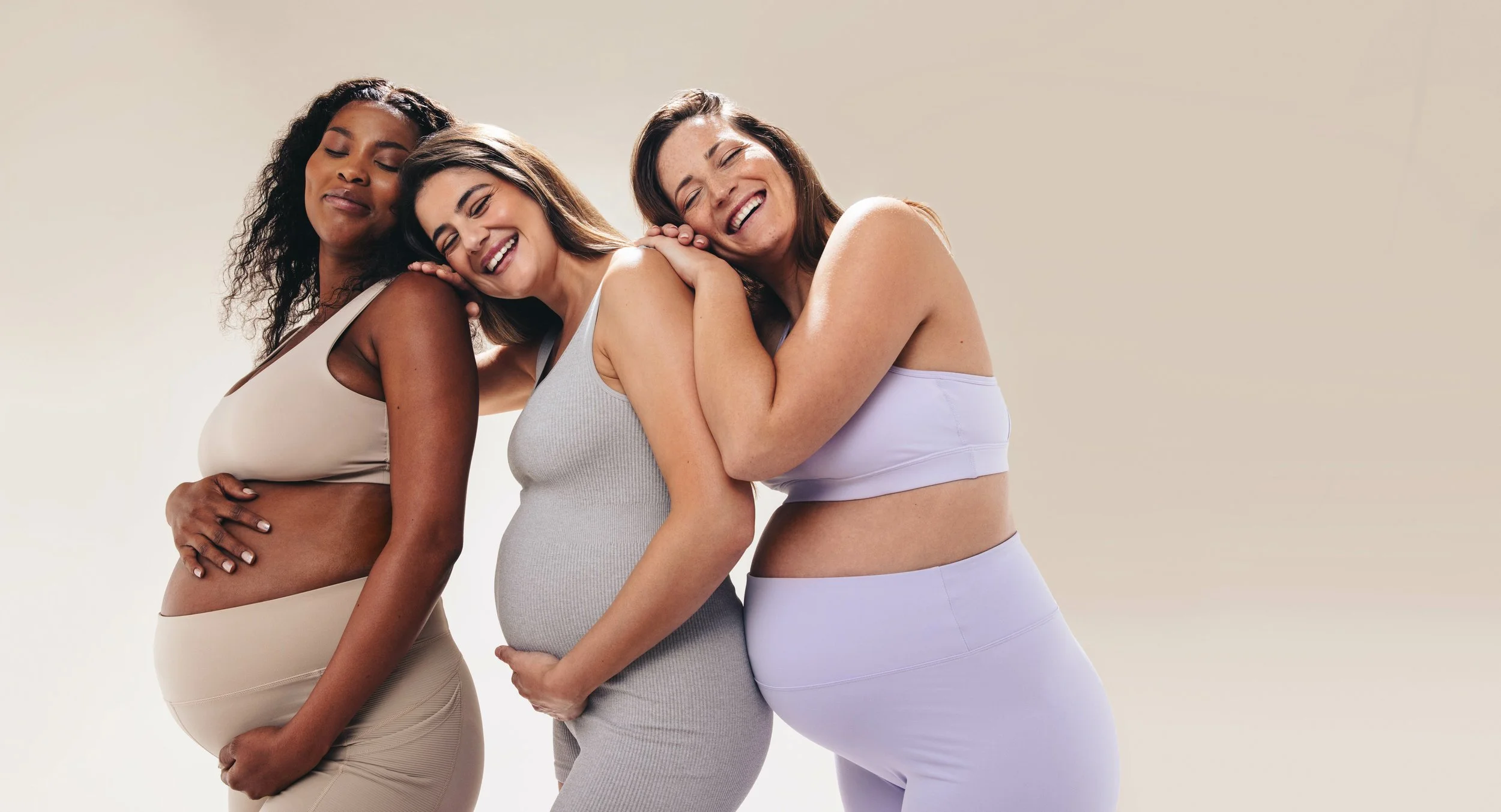 Three women with different skin tones, one pregnant, smiling and hugging each other, wearing light-colored comfortable clothing, standing close together against a neutral background.