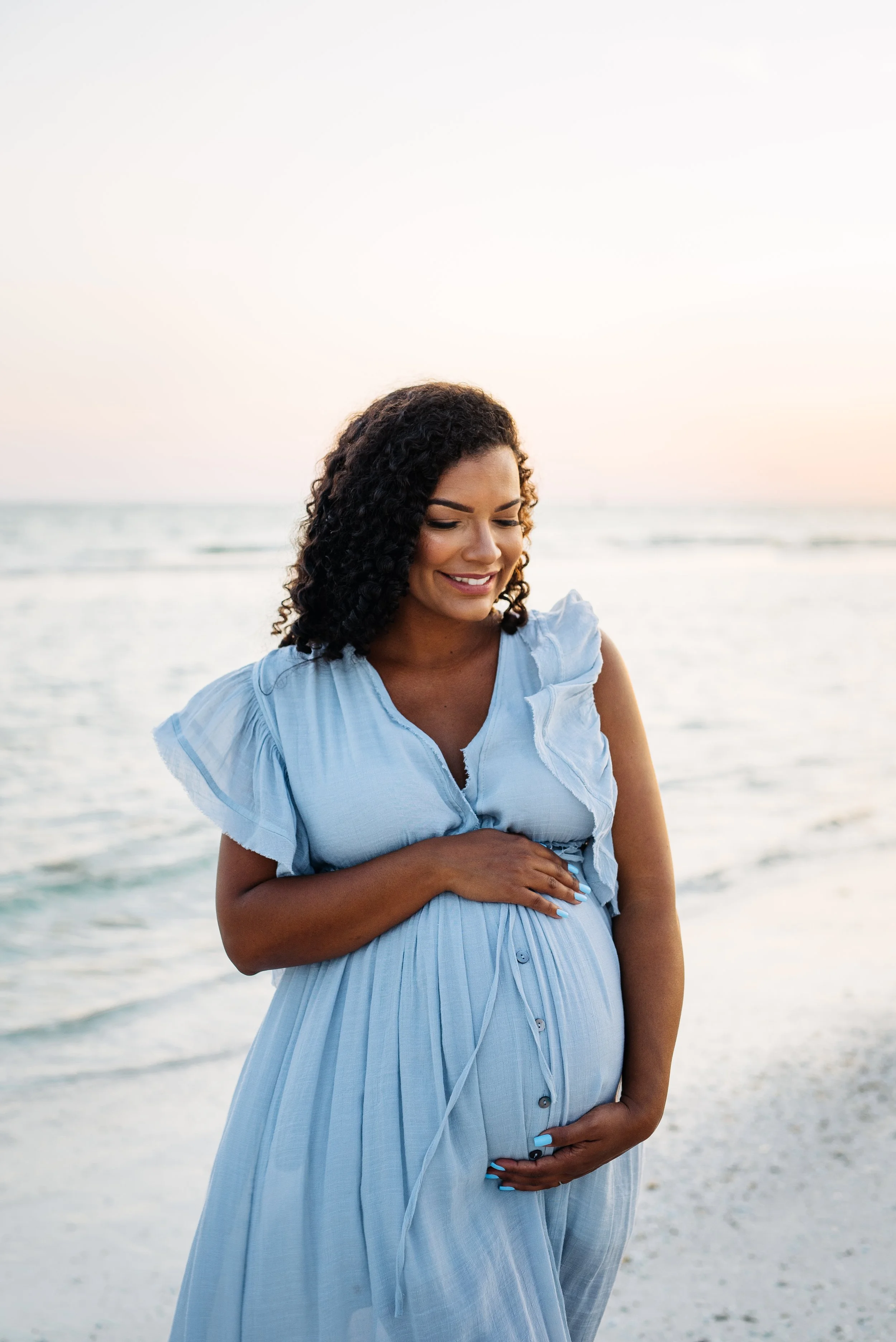 A pregnant woman in a light blue dress smiling at the beach at sunset, gently touching her belly.
