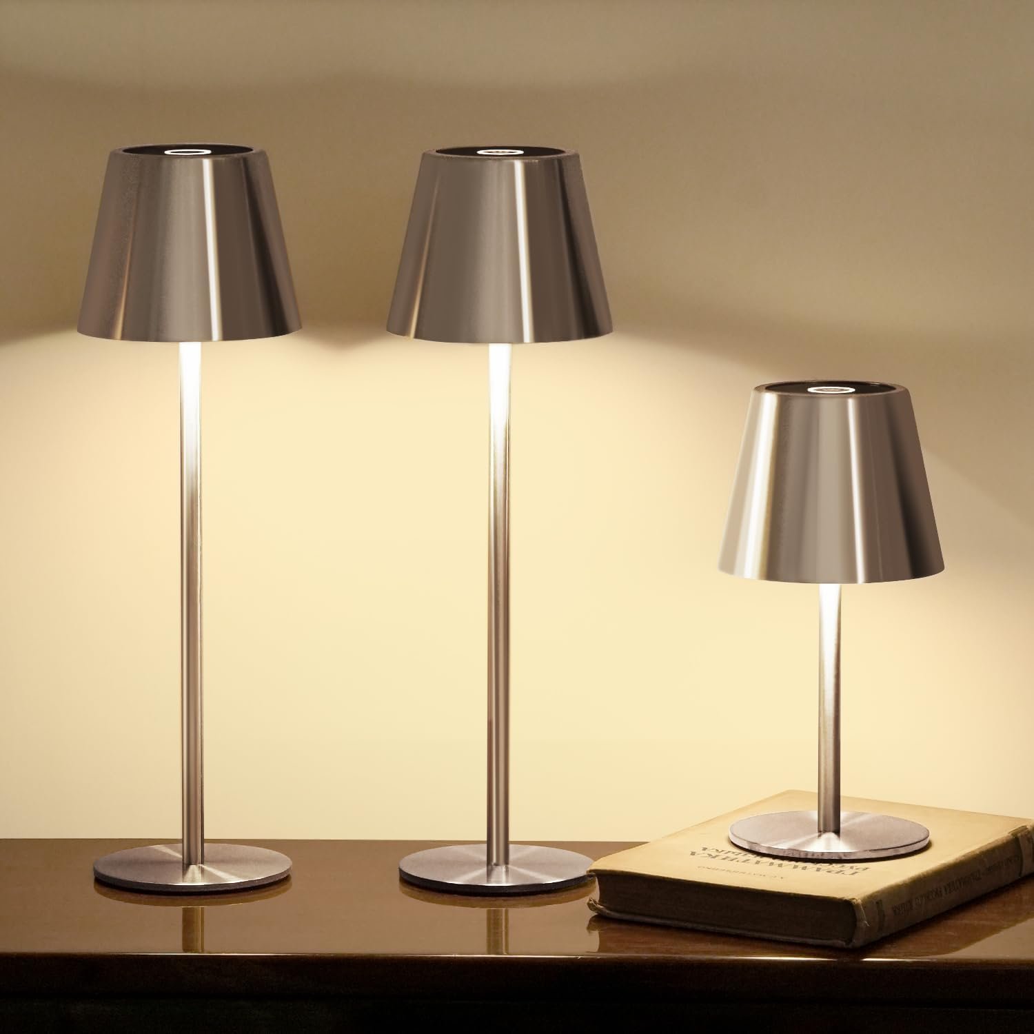 Three modern silver table lamps with conical shades, two illuminated and one off, placed on a dark polished surface with an old book underneath.