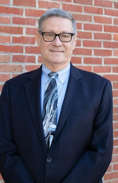 A middle-aged man with glasses, gray hair, wearing a dark suit, light blue shirt, and a patterned tie, standing in front of a brick wall.