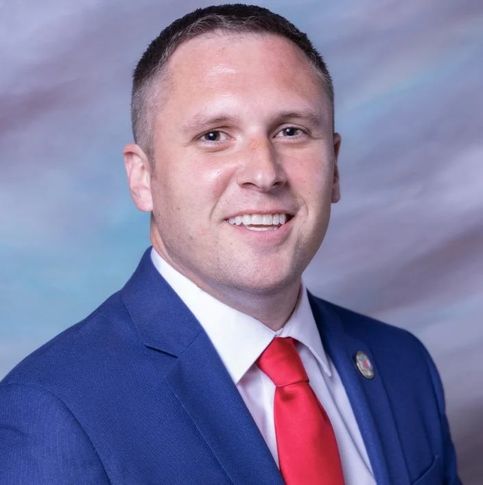 A man in a blue suit with a red tie smiling, against a cloudy sky background.