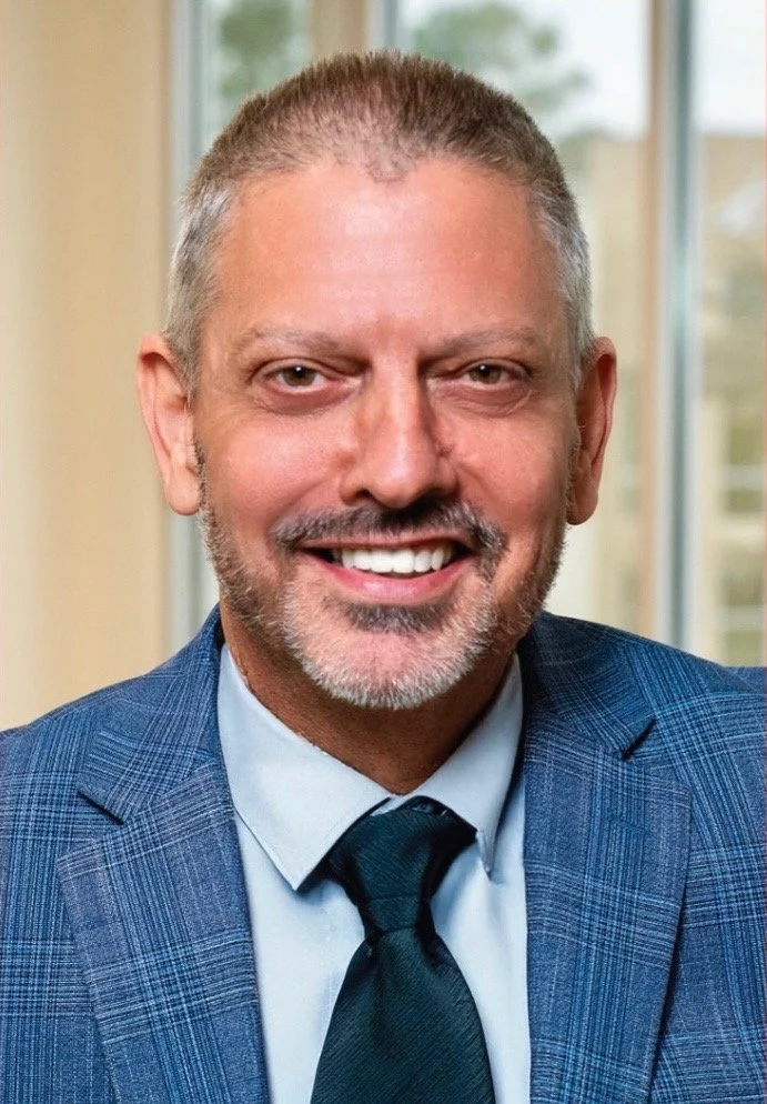 A smiling middle-aged man with short gray hair and a beard, wearing a blue checkered suit, light blue shirt, and dark tie, sitting indoors with a window in the background.