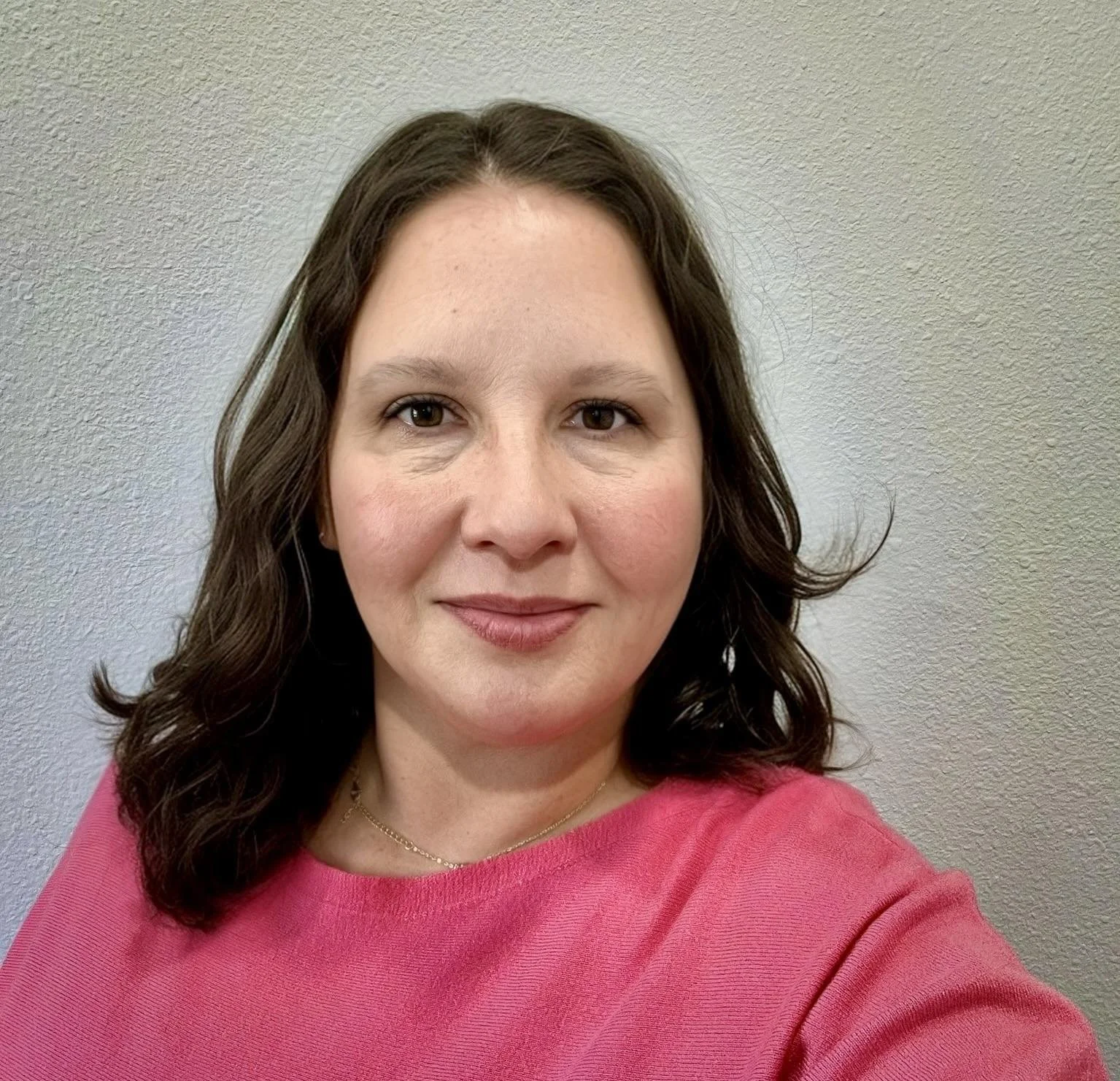 A woman with shoulder-length brown curly hair wearing a pink top stands against a plain beige textured wall.