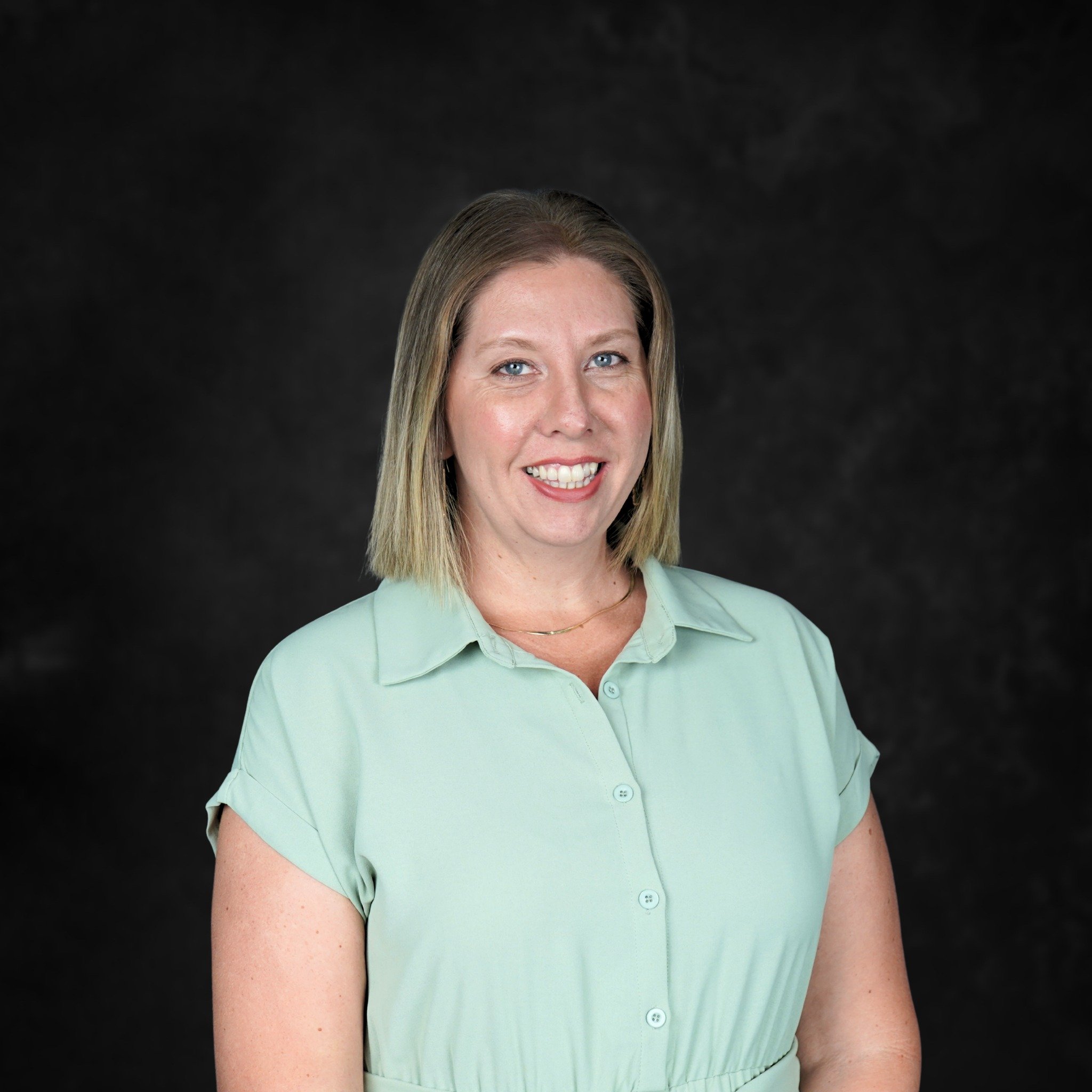 A woman with shoulder-length blonde hair wearing a light green short-sleeved button-up shirt smiling against a dark background.