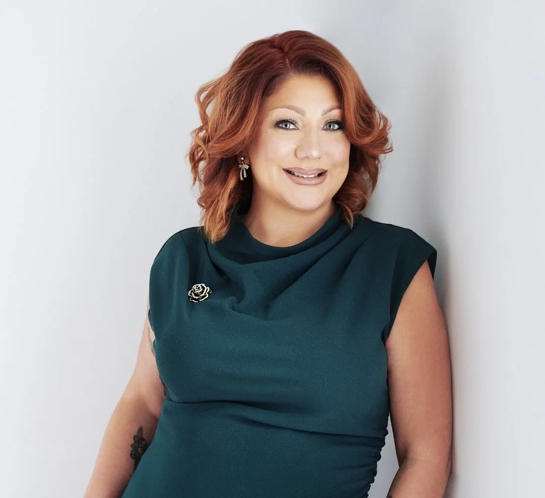 A woman with shoulder-length red hair, wearing a teal dress with a brooch, standing against a light gray background.