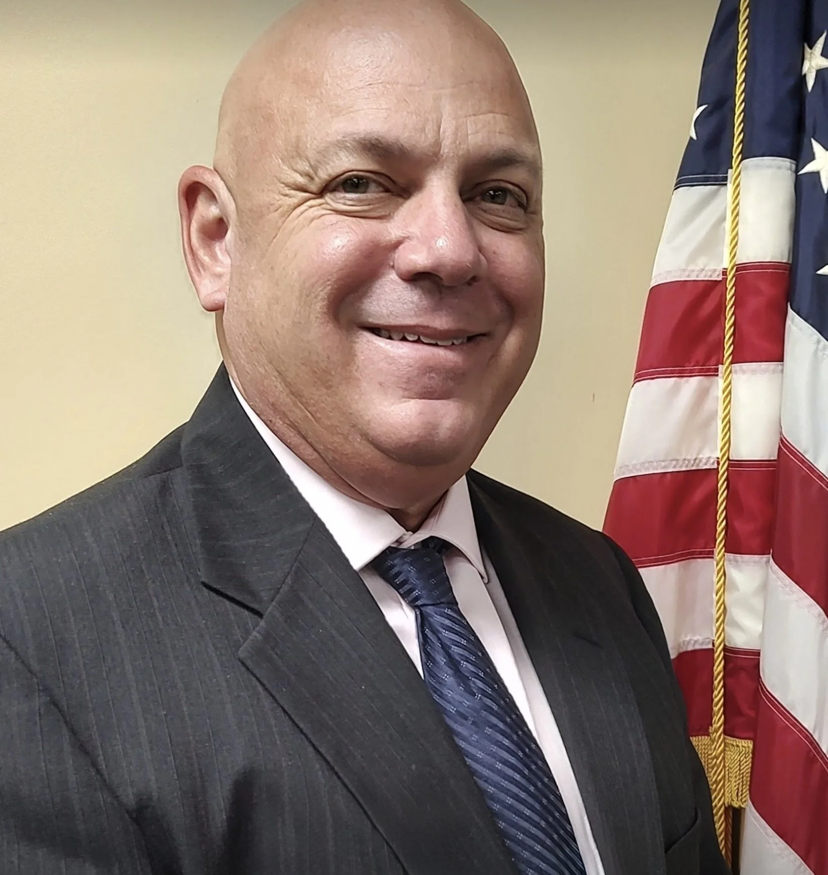 A smiling man in a gray suit, white shirt, and blue striped tie, standing next to an American flag.